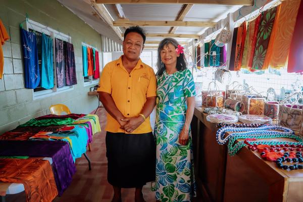 Two people stand amongst some clothing and handicrafts in a market stall and smile for the camera