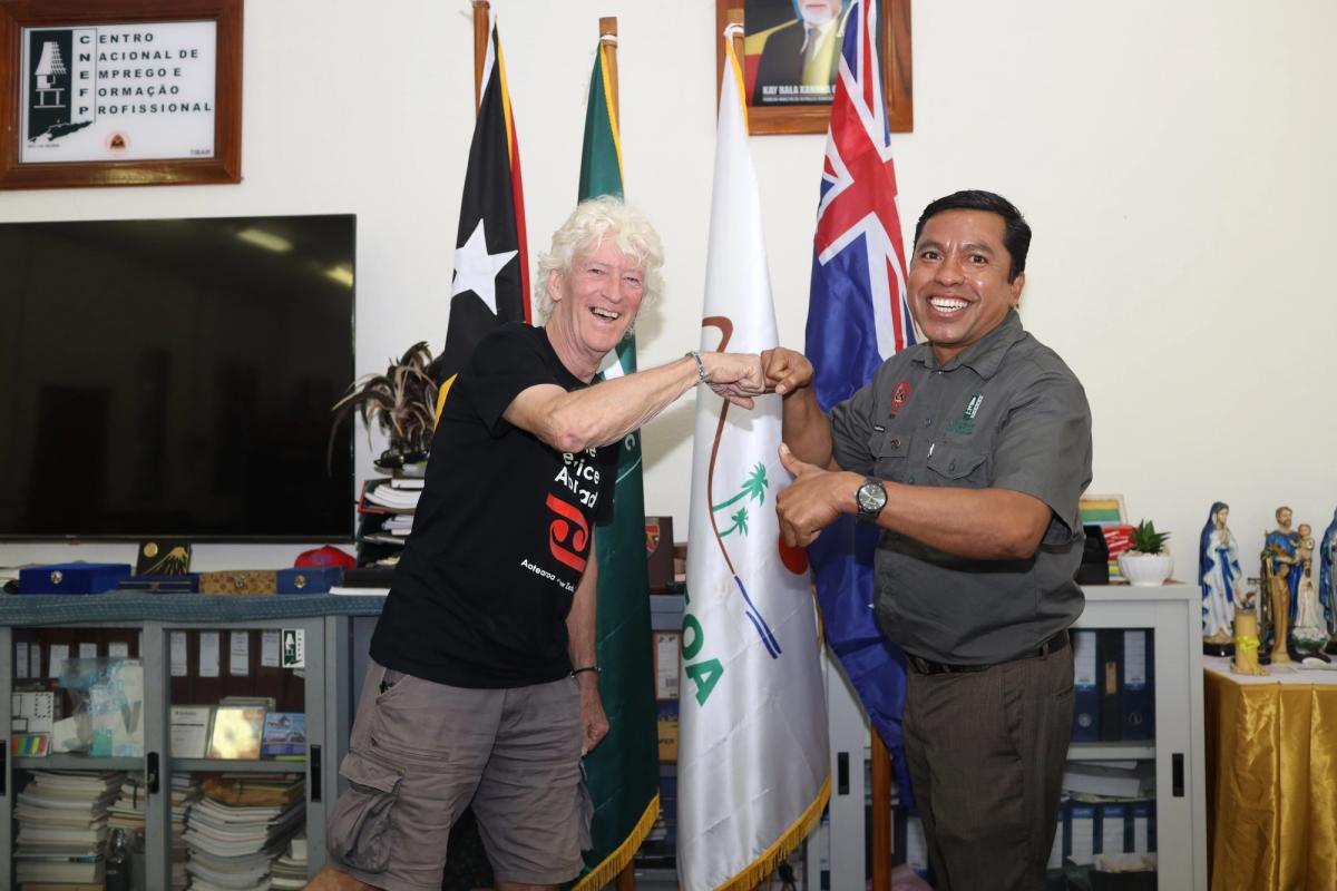 Two men smiling and doing a fist bump in a room in front of flags.