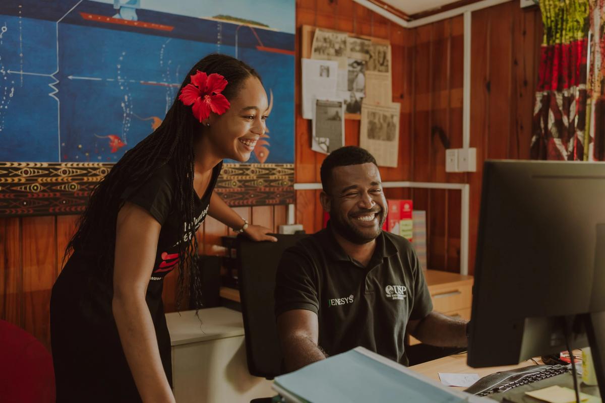 Two people smiling and working at a desk on a computer.