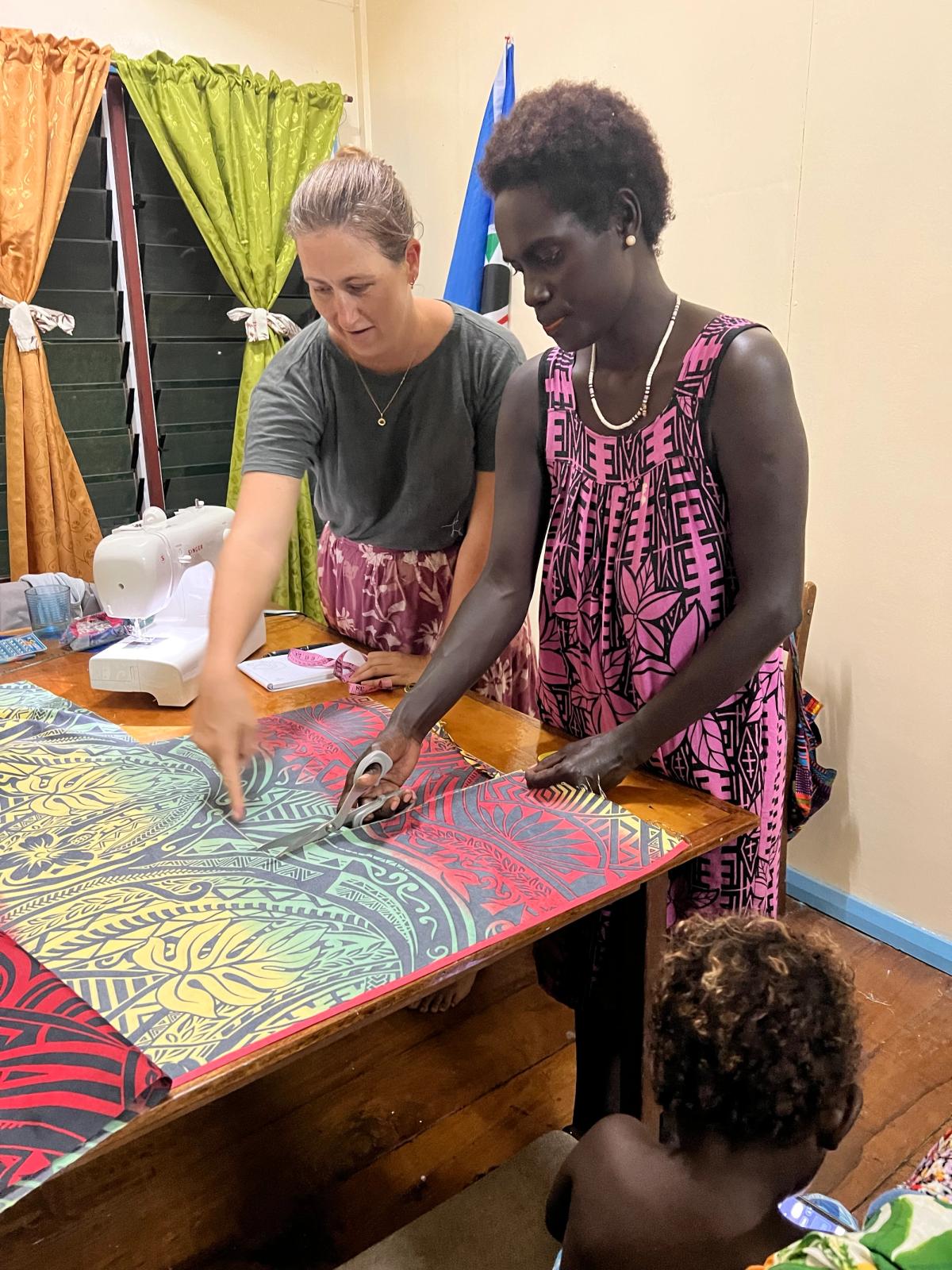 One woman cutting a piece of fabric while another woman guides her One woman cutting a piece of fabric while another woman guides her