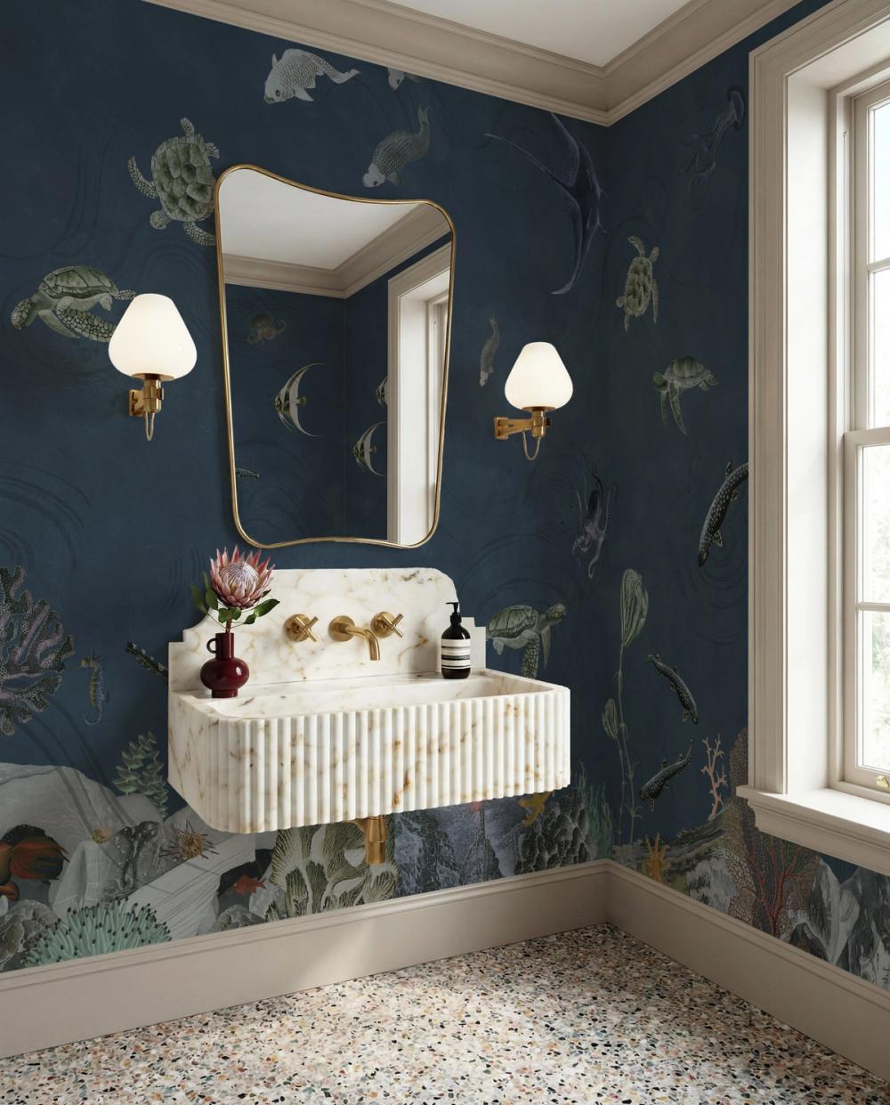 A photo of a poder room with marble sink, a mirror with brass frame and a undersea themed dark blue wallpaper on the walls