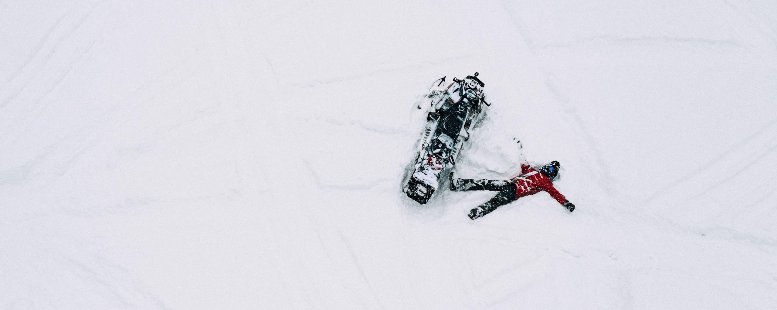 Aeriel photo of man laying in the snow next to snowmobile in Aspen