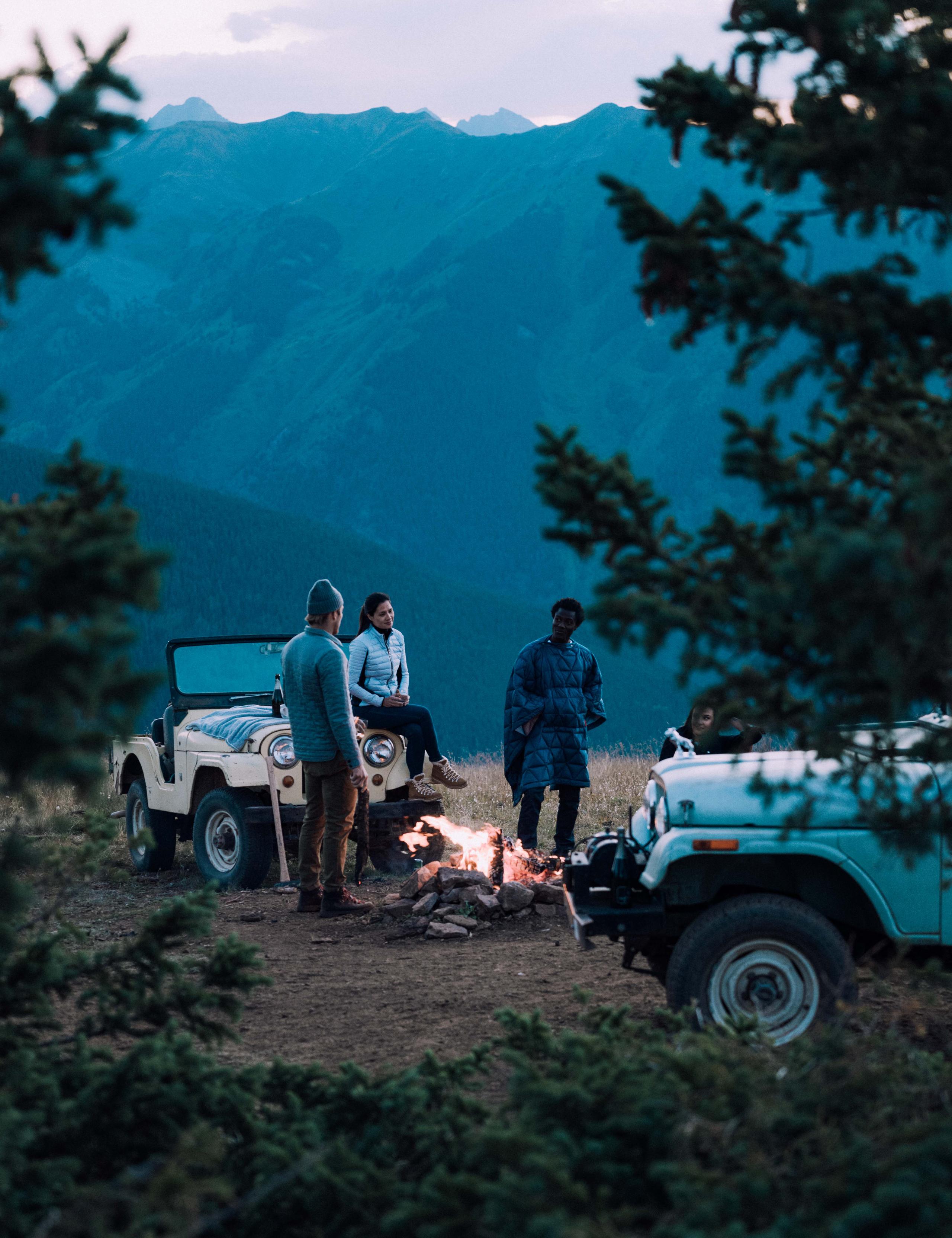 Two men and one woman tending to a fire at dusk next to two vintage Jeeps on mountain in Aspen
