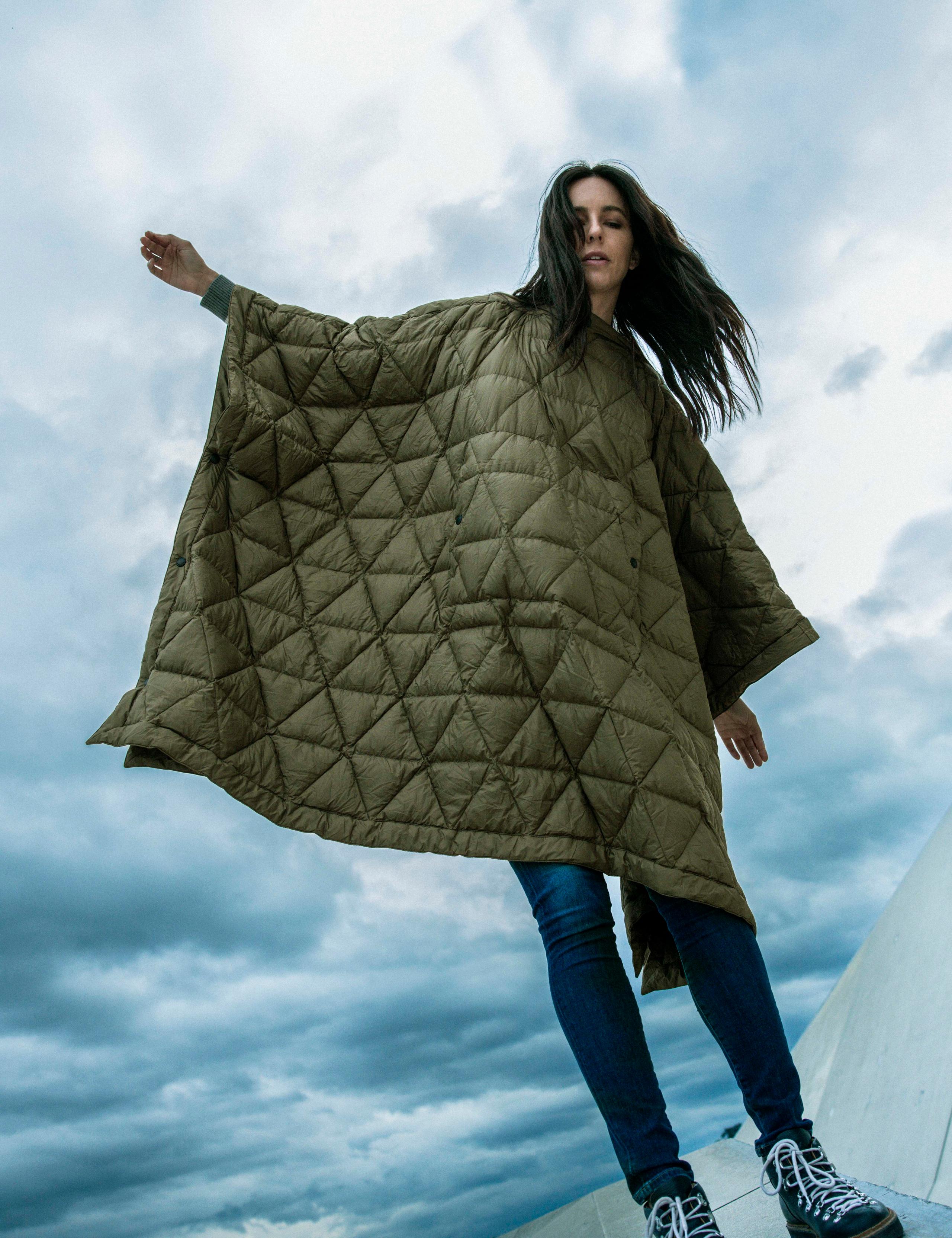 Woman wearing Down Poncho with arms out against blue sky and clouds