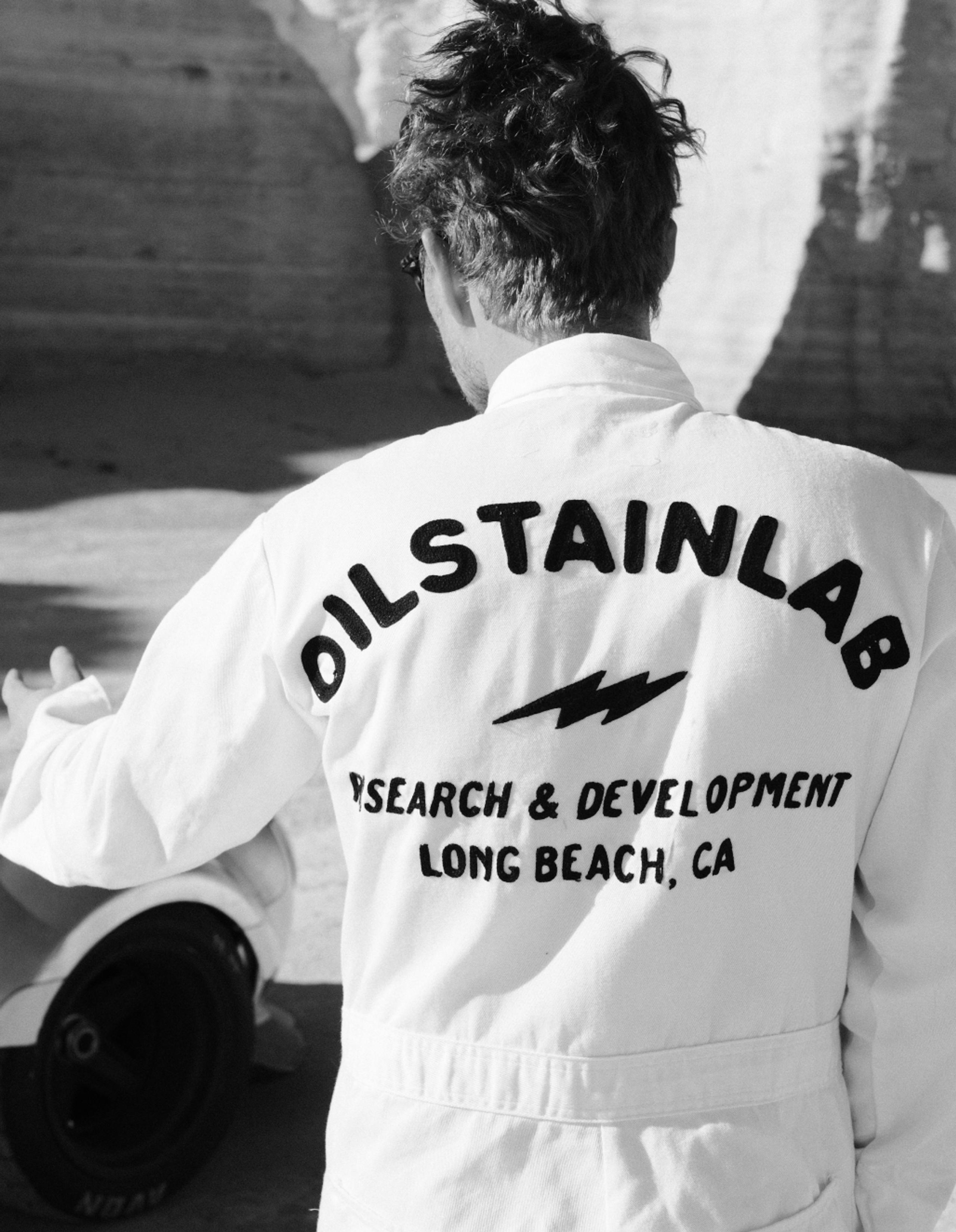 Man wearing race jumpsuit with lettering that says OILSTAINLAB - Research & Development, Long Beach CA