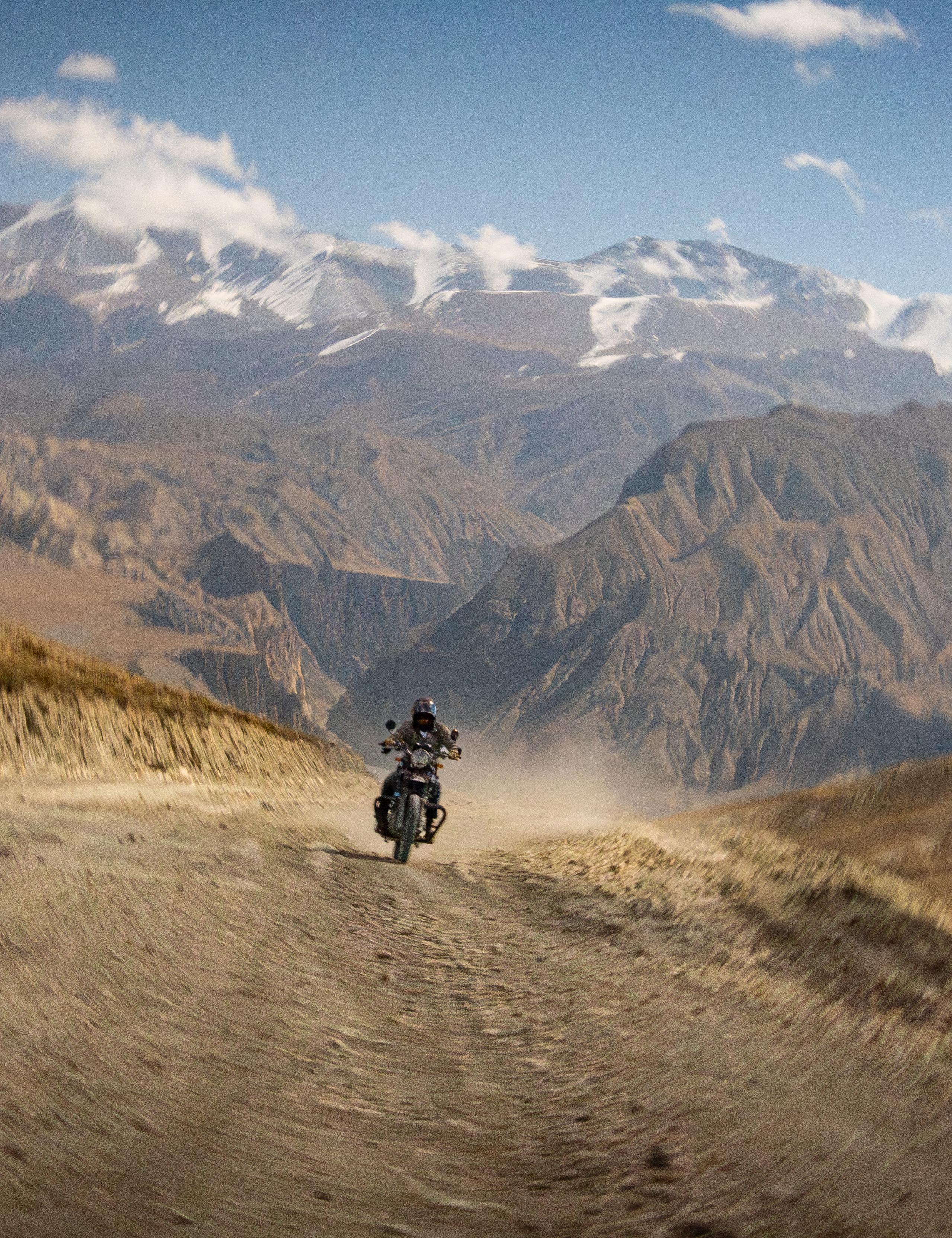 Motorcyclist riding and kicking up dust on dirt road with Himalayan mountains in the background