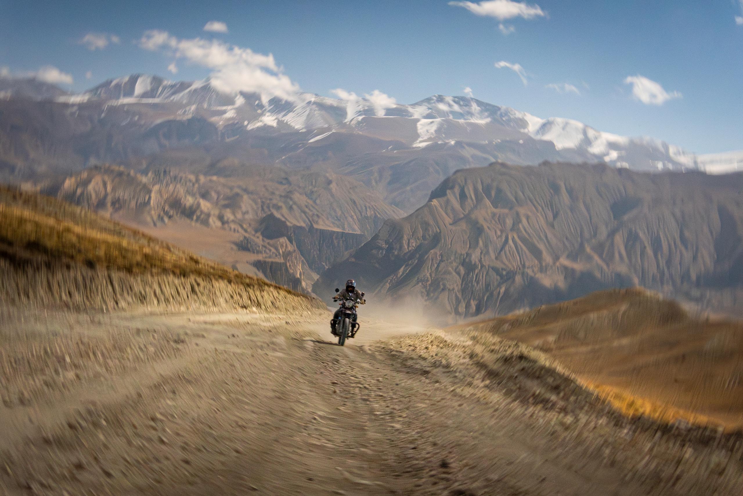 Motorcyclist riding and kicking up dust on dirt road with Himalayan mountains in the background