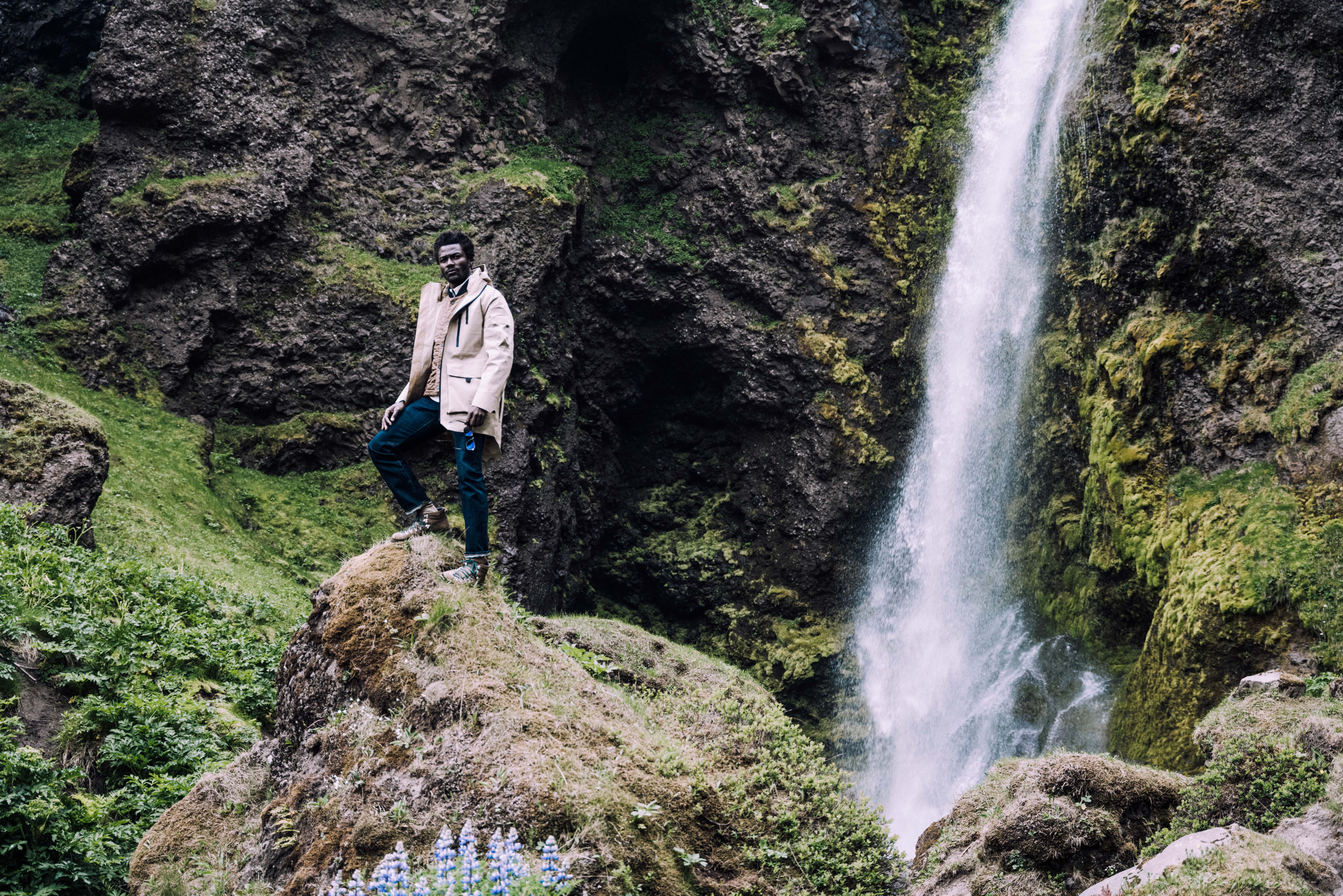 Man wearing AETHER + Mackintosh Field Parka on rocky terrain next to waterfall in Iceland
