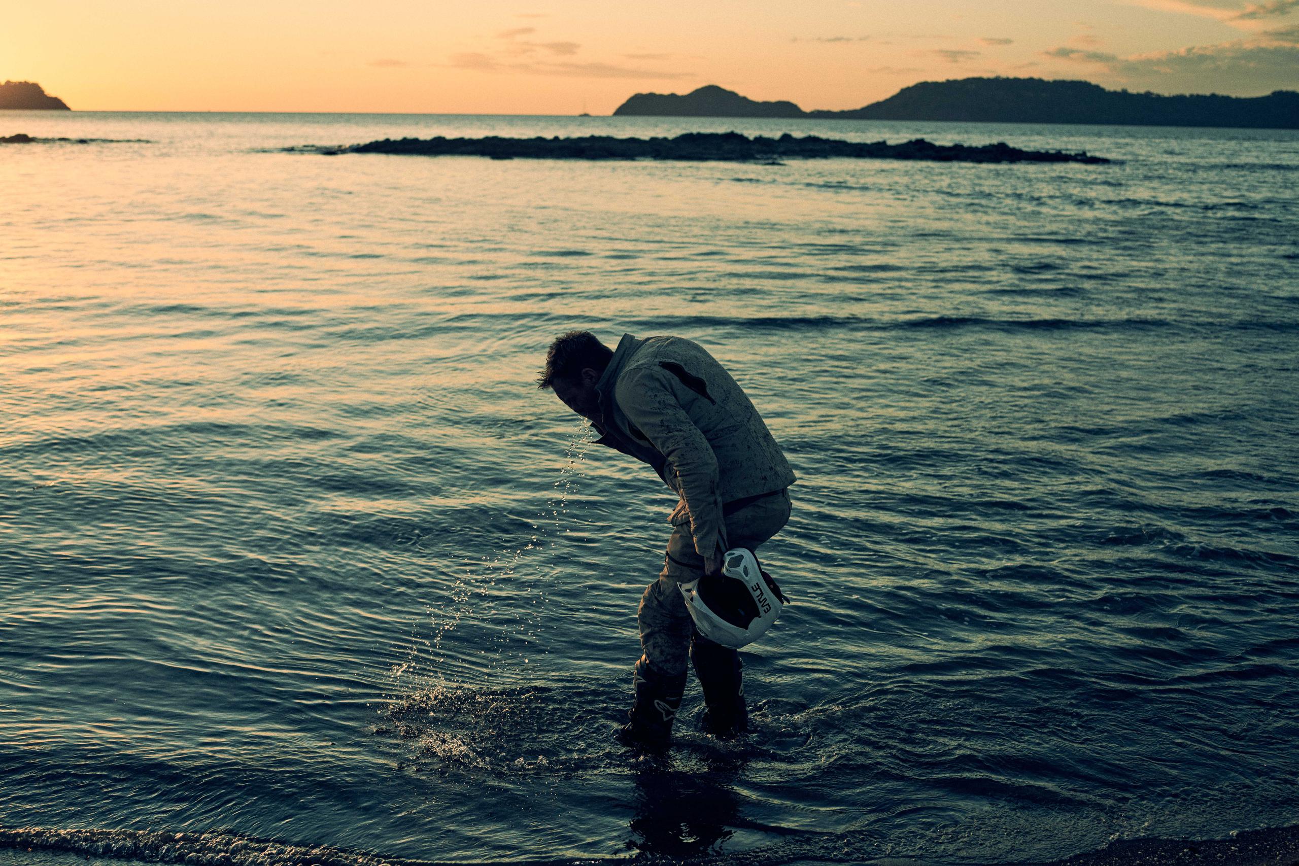 Alex Earle in motorcycle uniform at sunset standing in ocean off Costa Rica splashing water in face