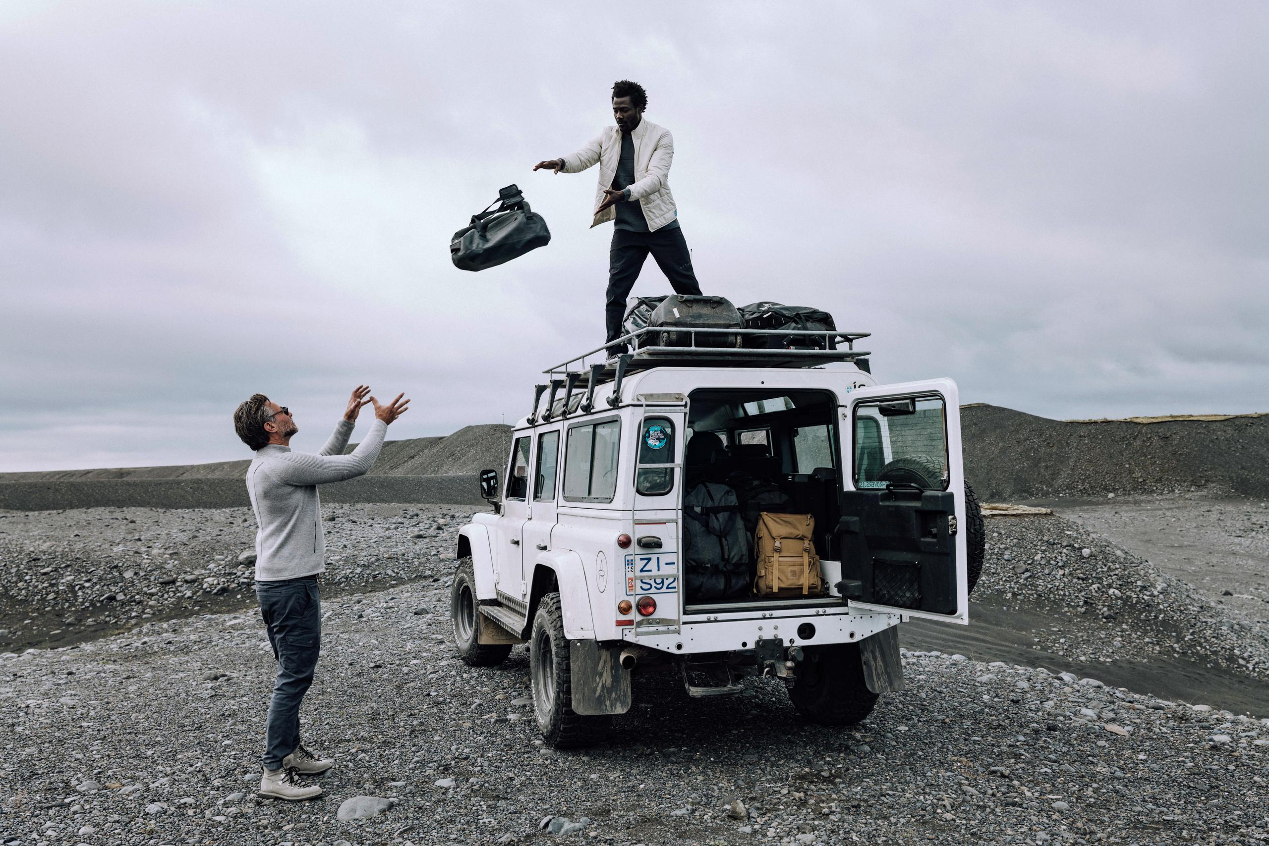 Man throwing luggage from top of white Defender to man below in Iceland landscape