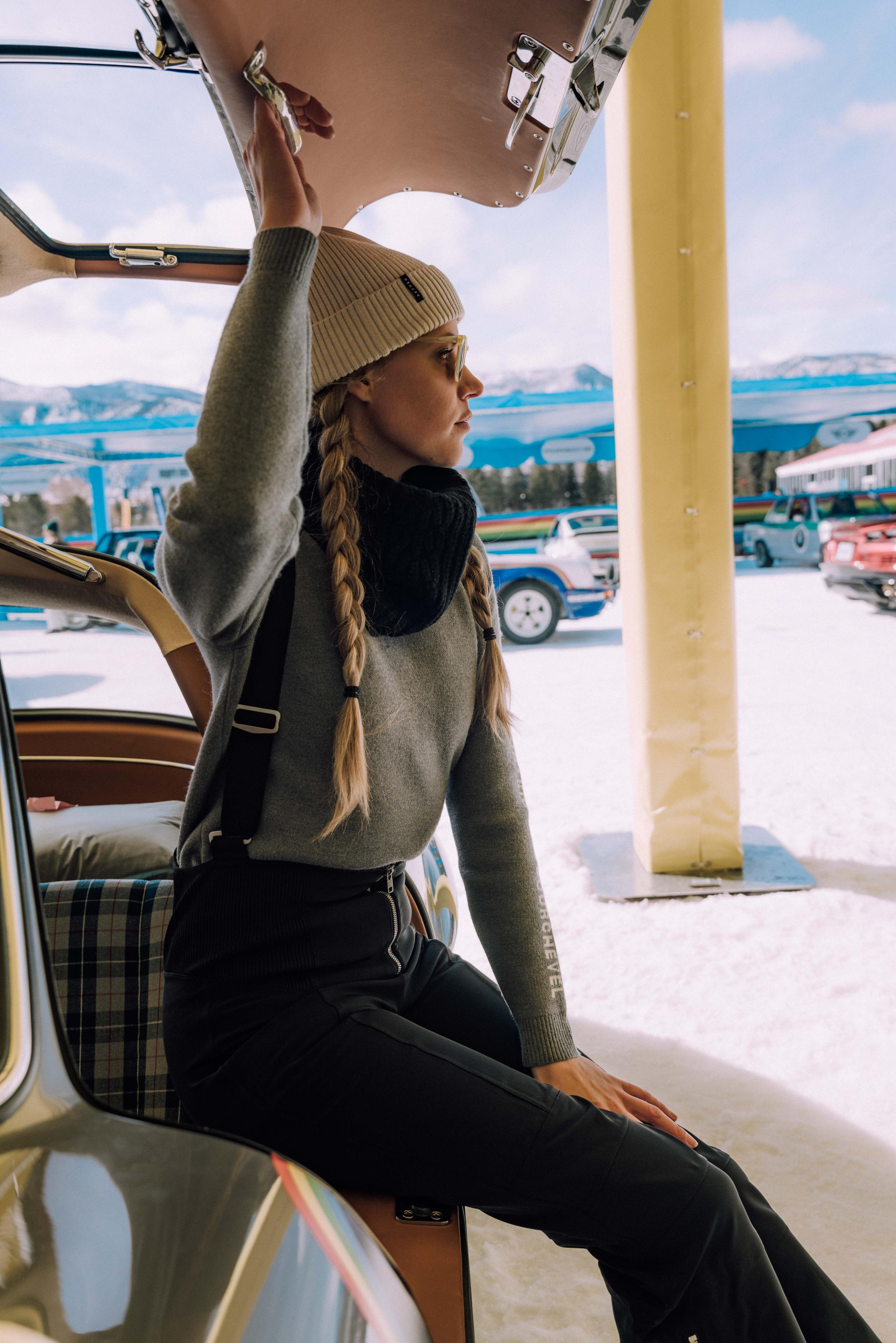 Woman wearing Aether sweater and snow bib sitting on back of vintage car