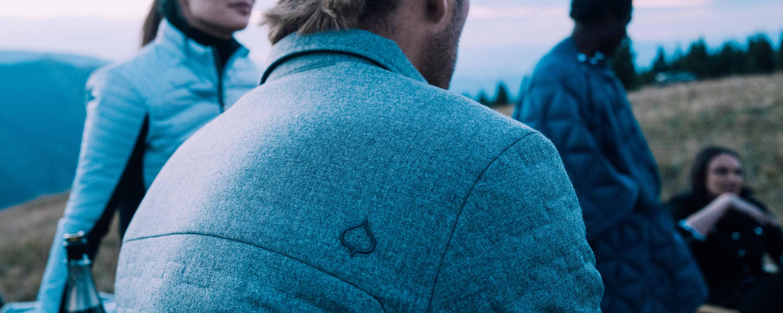 Closeup of man's shoulder with embroidered AspenX leaf logo on shirt with friends and campfire in the background