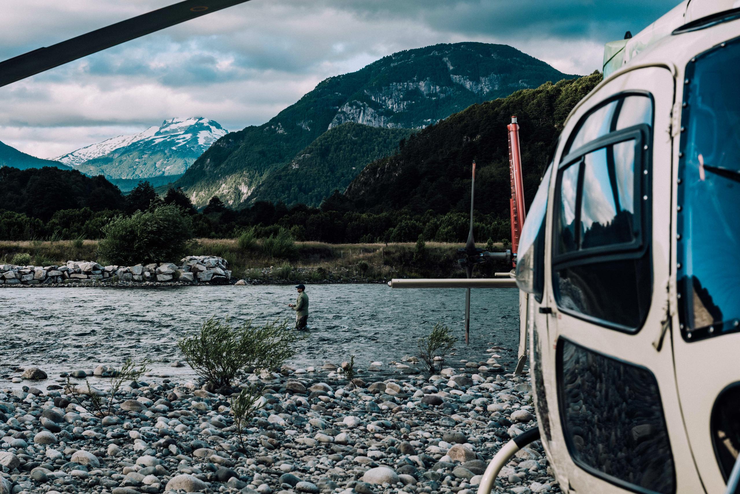 Man in river fly fishing with helicopter on the shore in the foreground and the mountains in the background