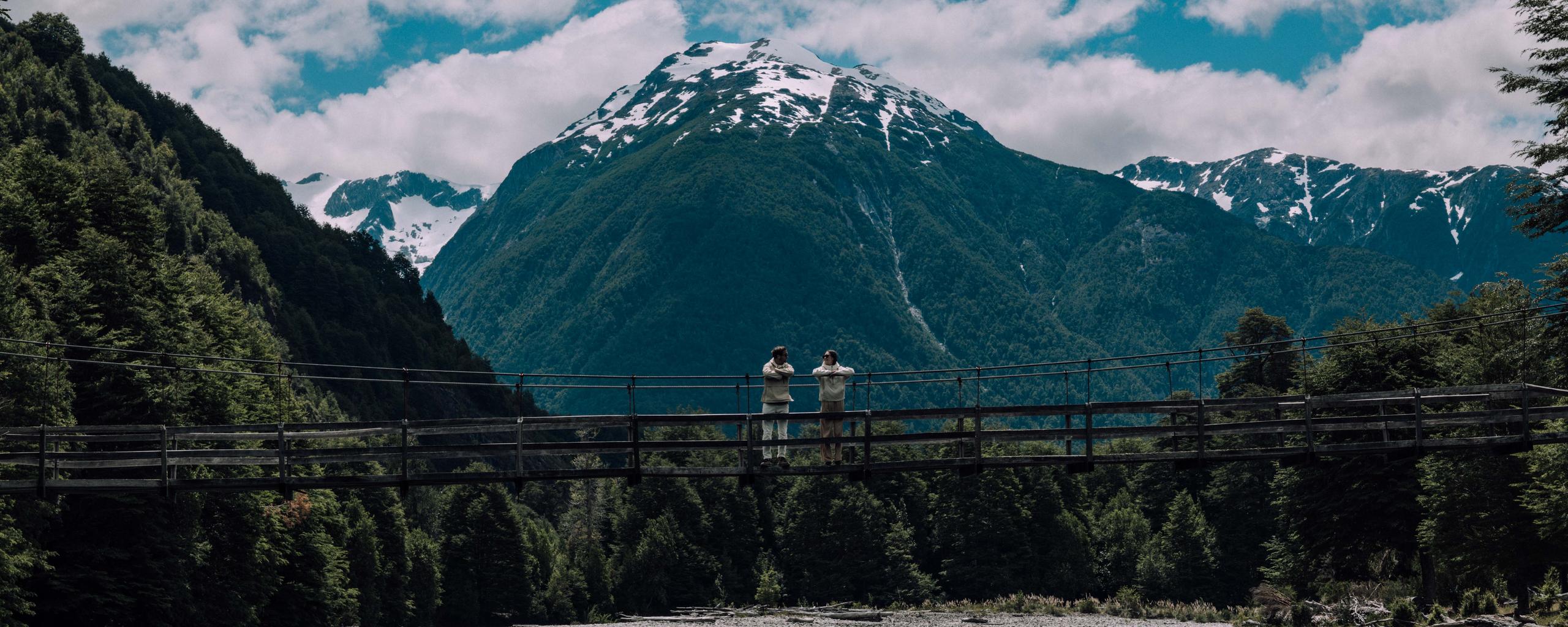Man and woman standing on wood bridge with mountains of Patagonia in background