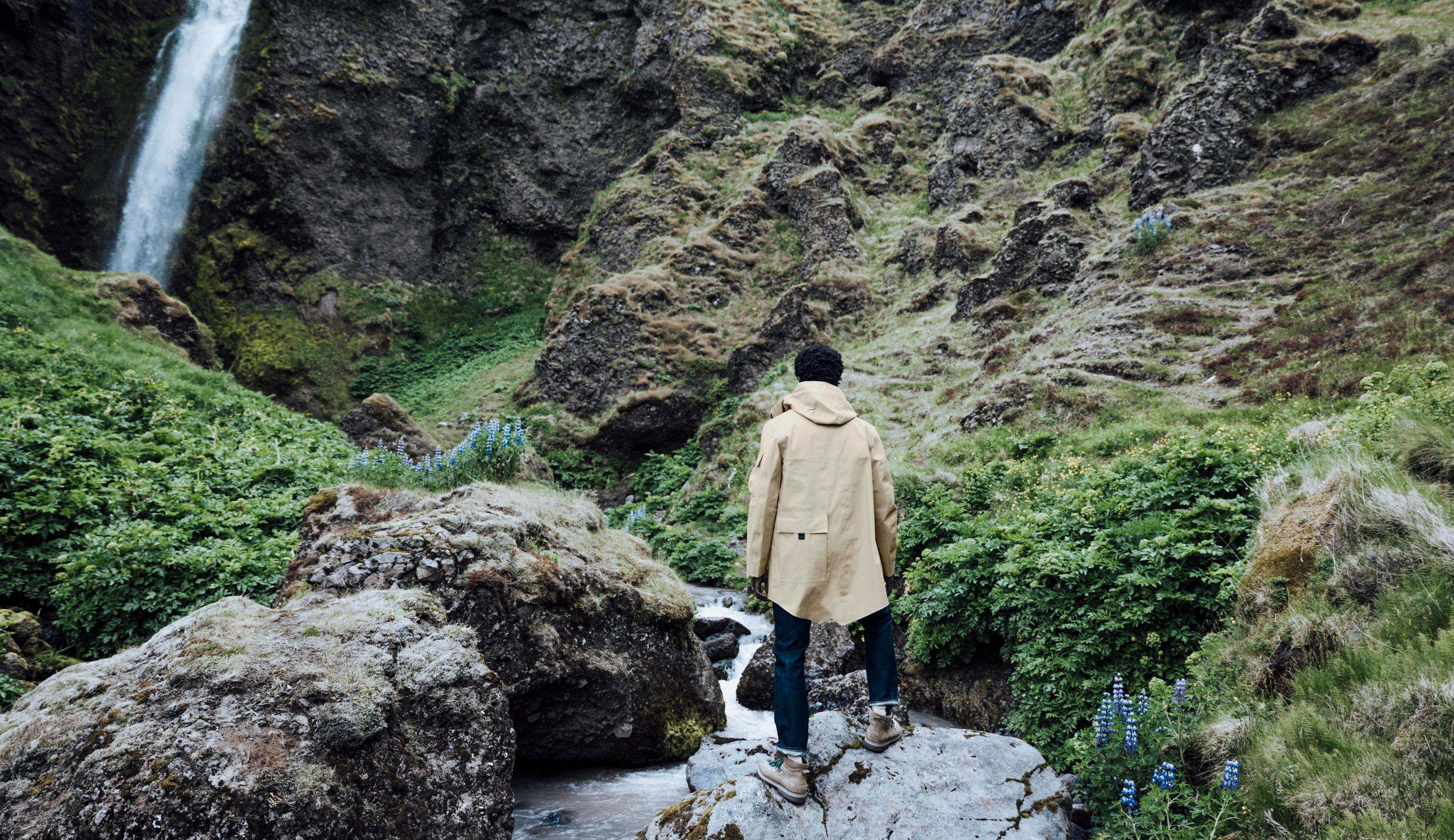 Man in AETHER + Mackintosh Field Parka standing in front of waterfall in Iceland. 