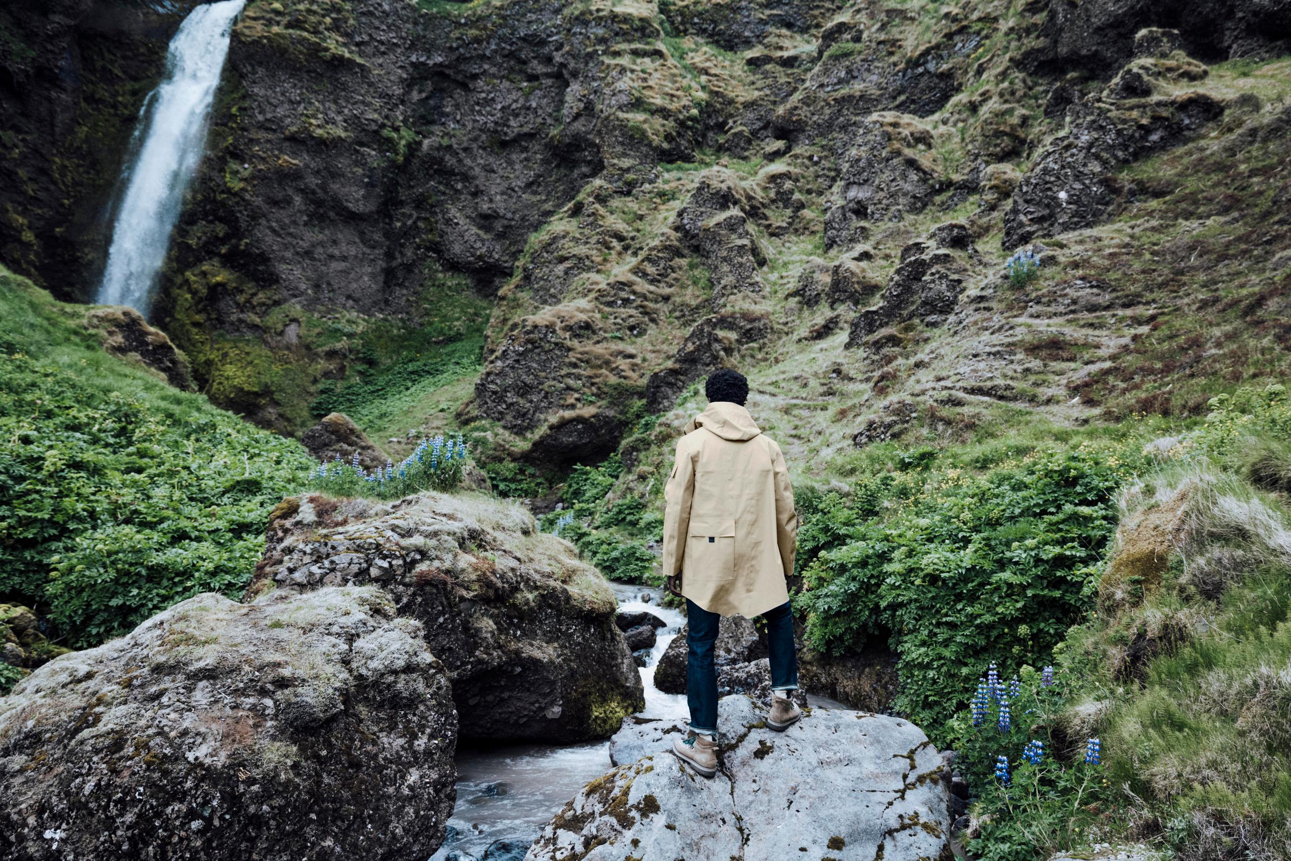 Man in AETHER + Mackintosh jacket hiking on rocky terrain next to waterfall in Iceland