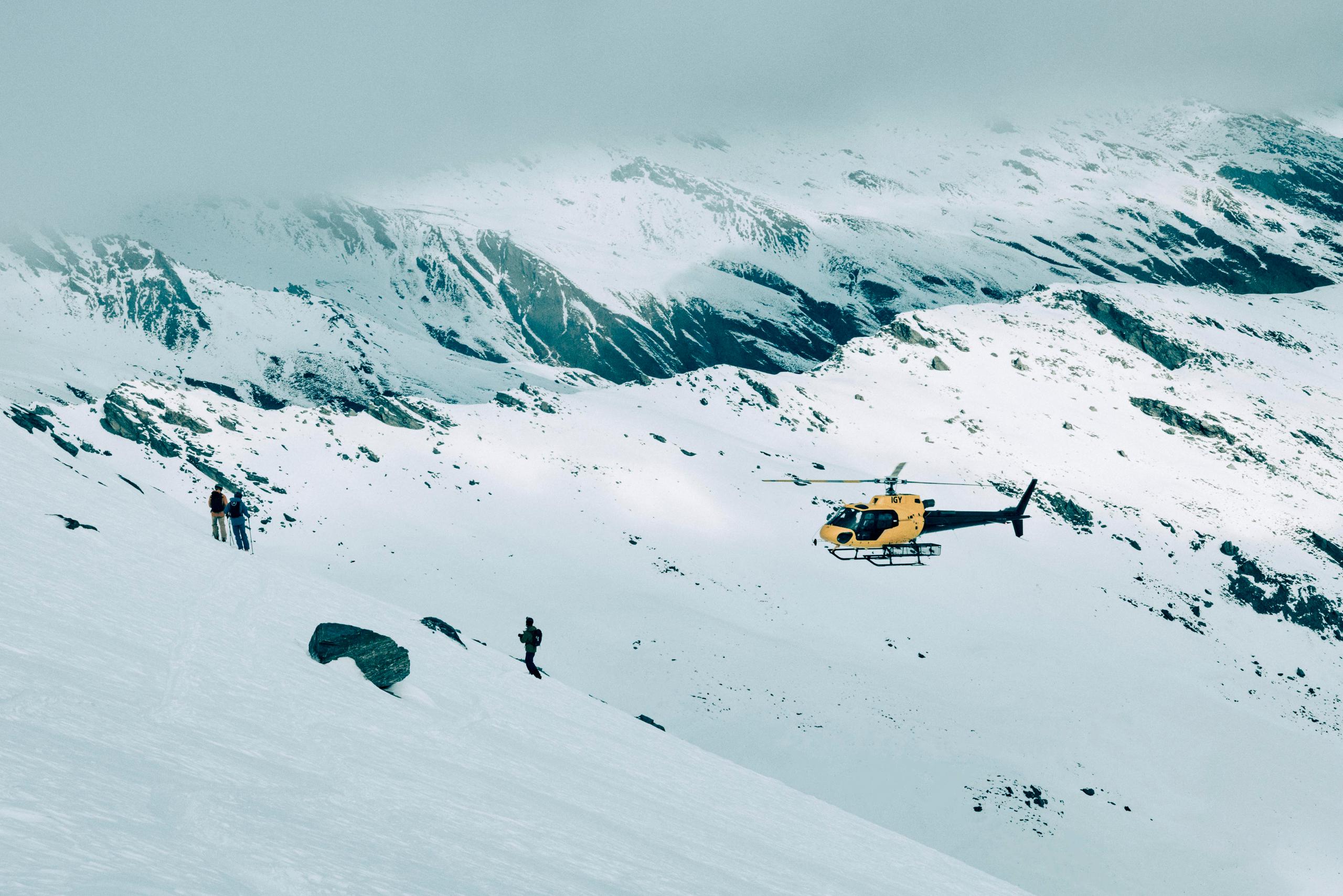 Helicopter is flying over a few skiers and snowboarders on rocky and snowy New Zealand mountain. 