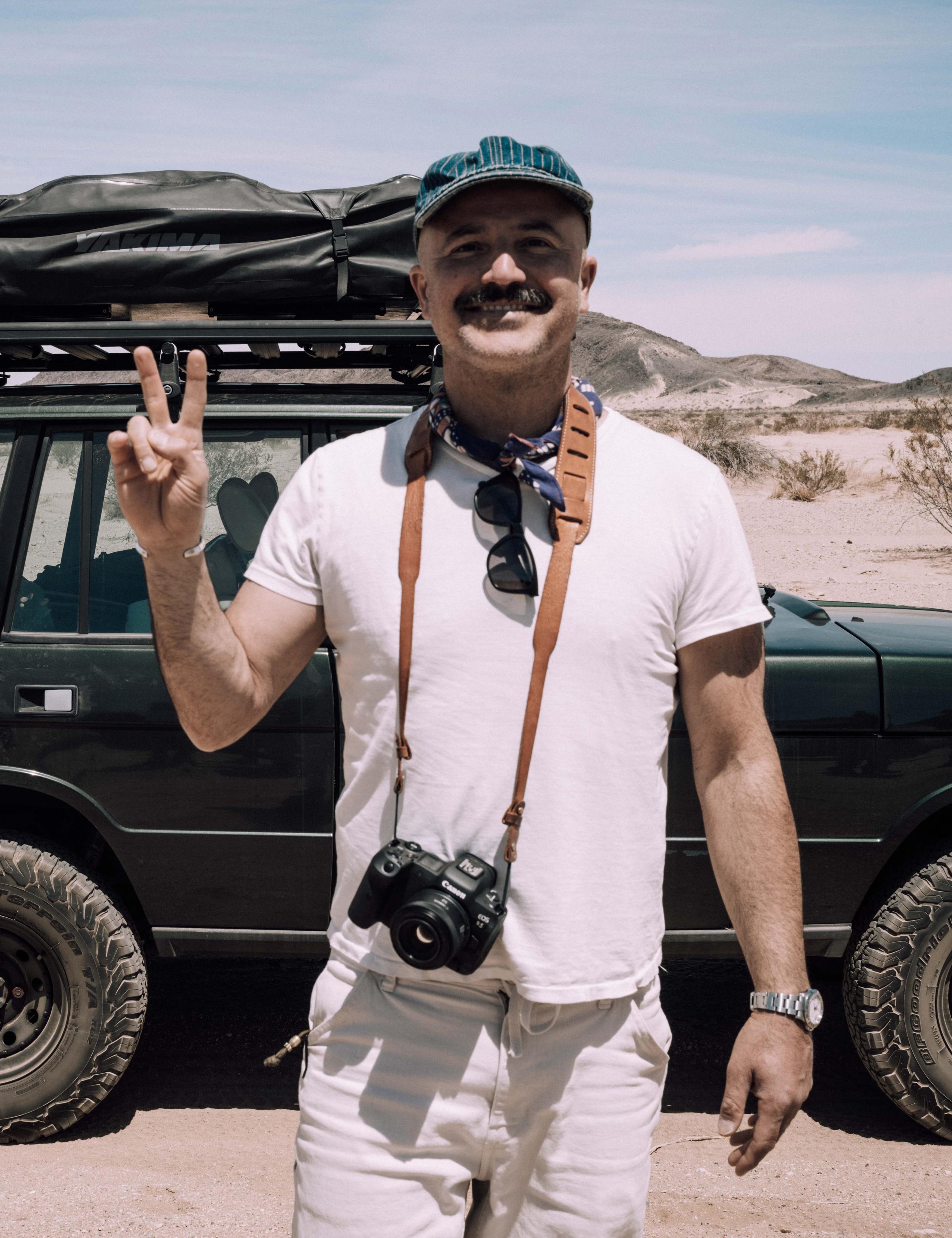 Man with camera around his neck giving the peace sign standing in front of Land Rover in Joshua Tree