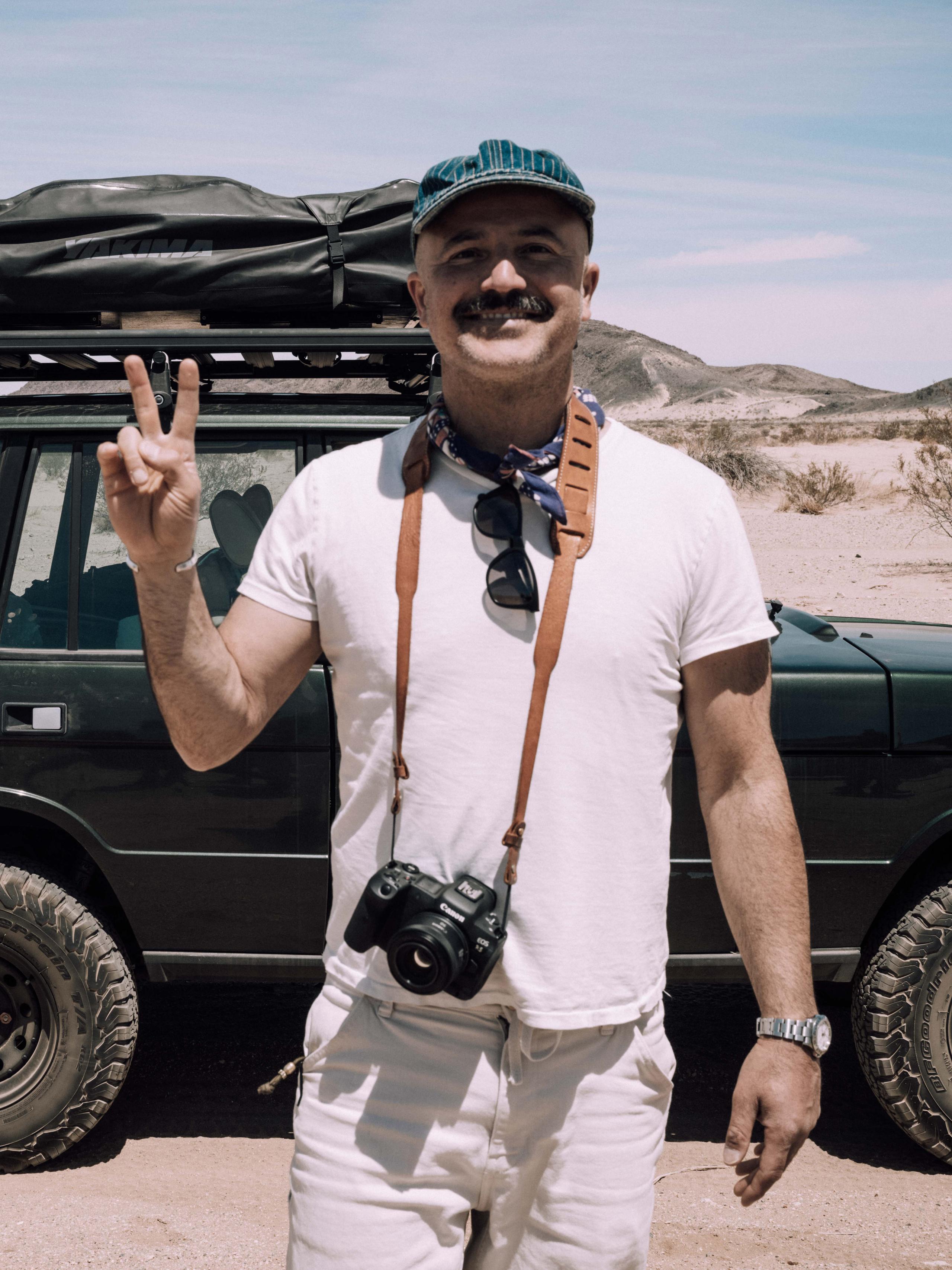 Man smiling and giving the peace sign, standing in front of vintage Land Rover in front of desert landscape