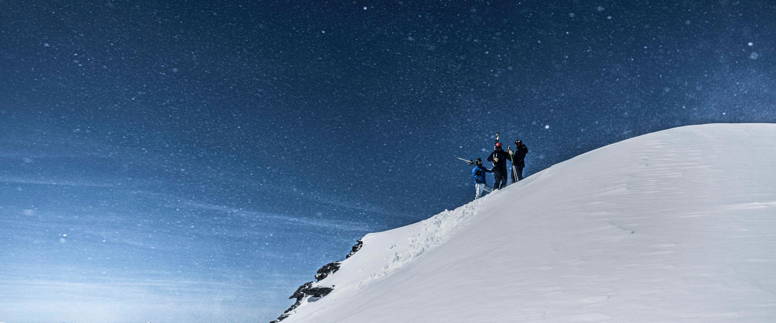 Three people standing with skis on a snowy mountain with a blue starry sky in the background