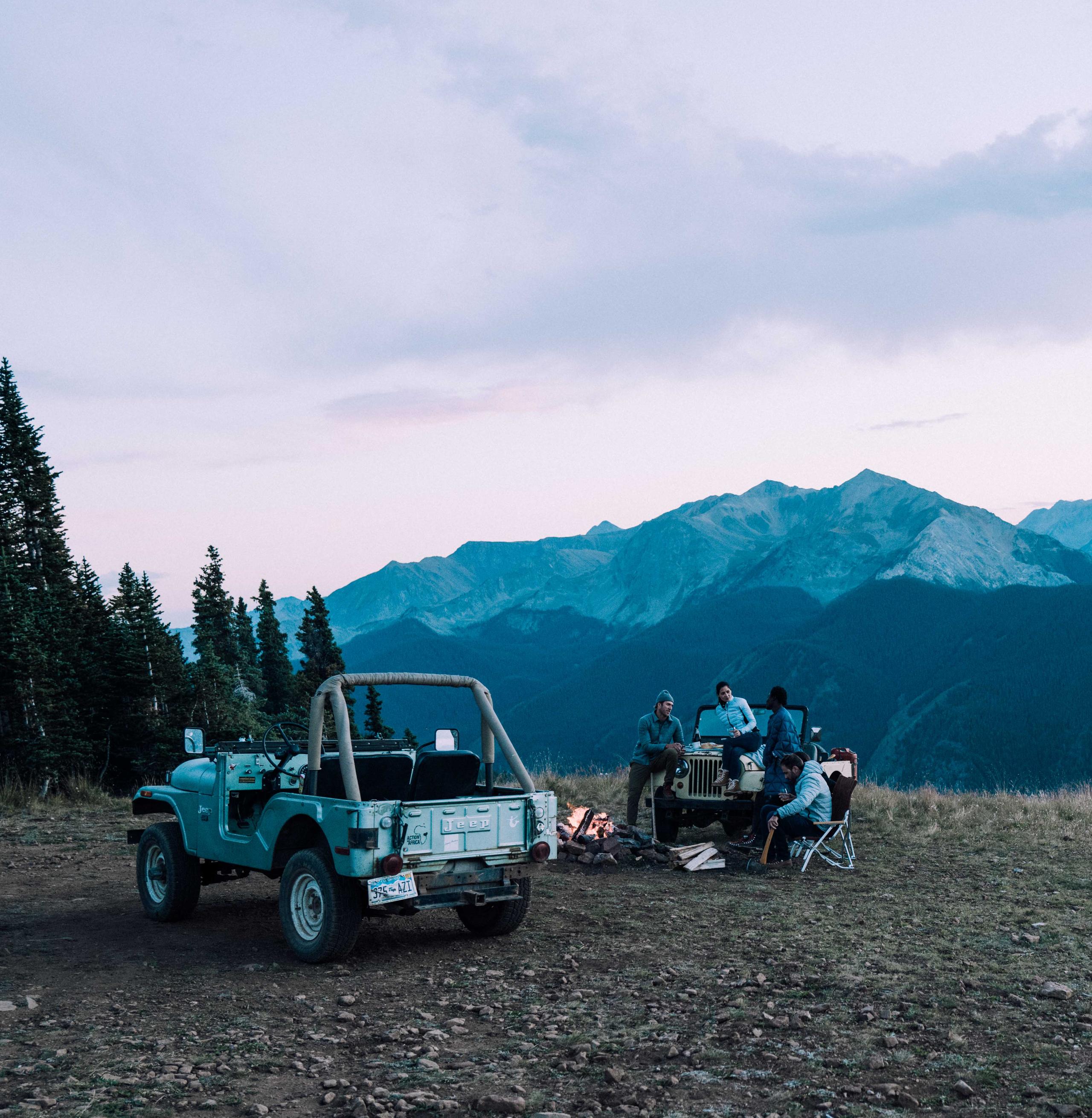 Group of people sitting next campfire and vintage Jeeps at sunset in the Aspen mountains