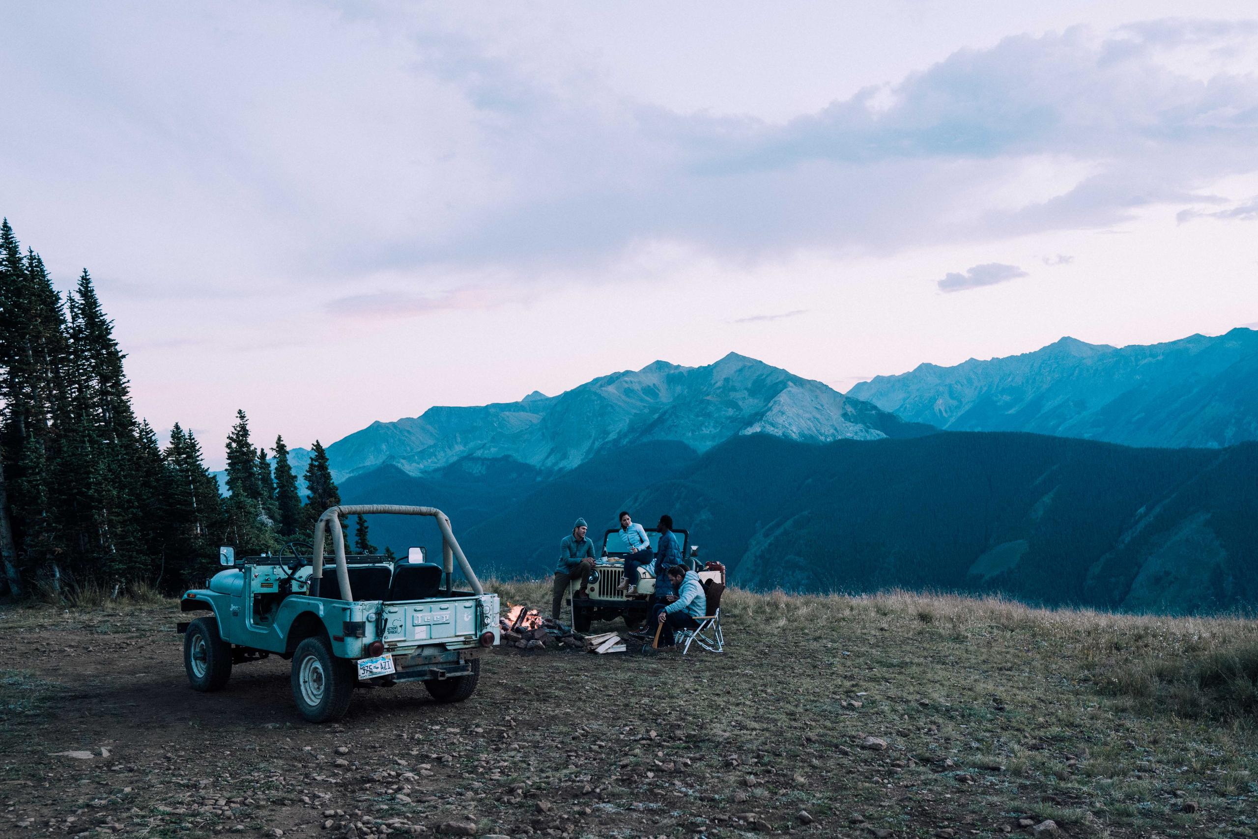Four friends around campfire and vintage Jeep at dusk in Aspen mountains