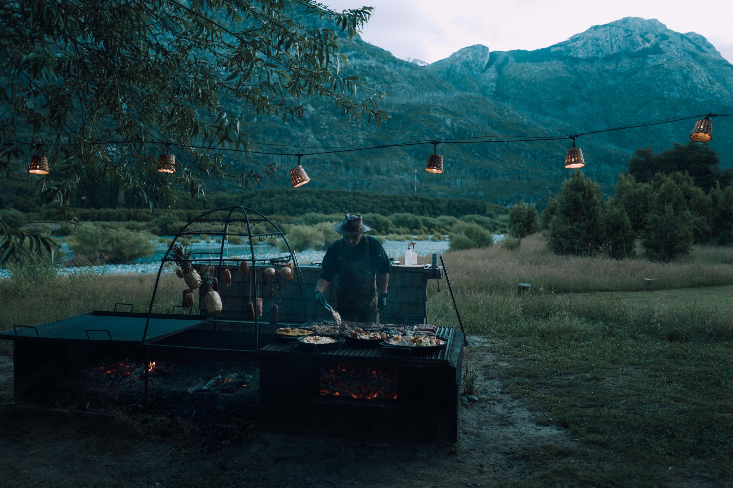 Man cooking on outdoor grill at dusk in front of river in Patagonia