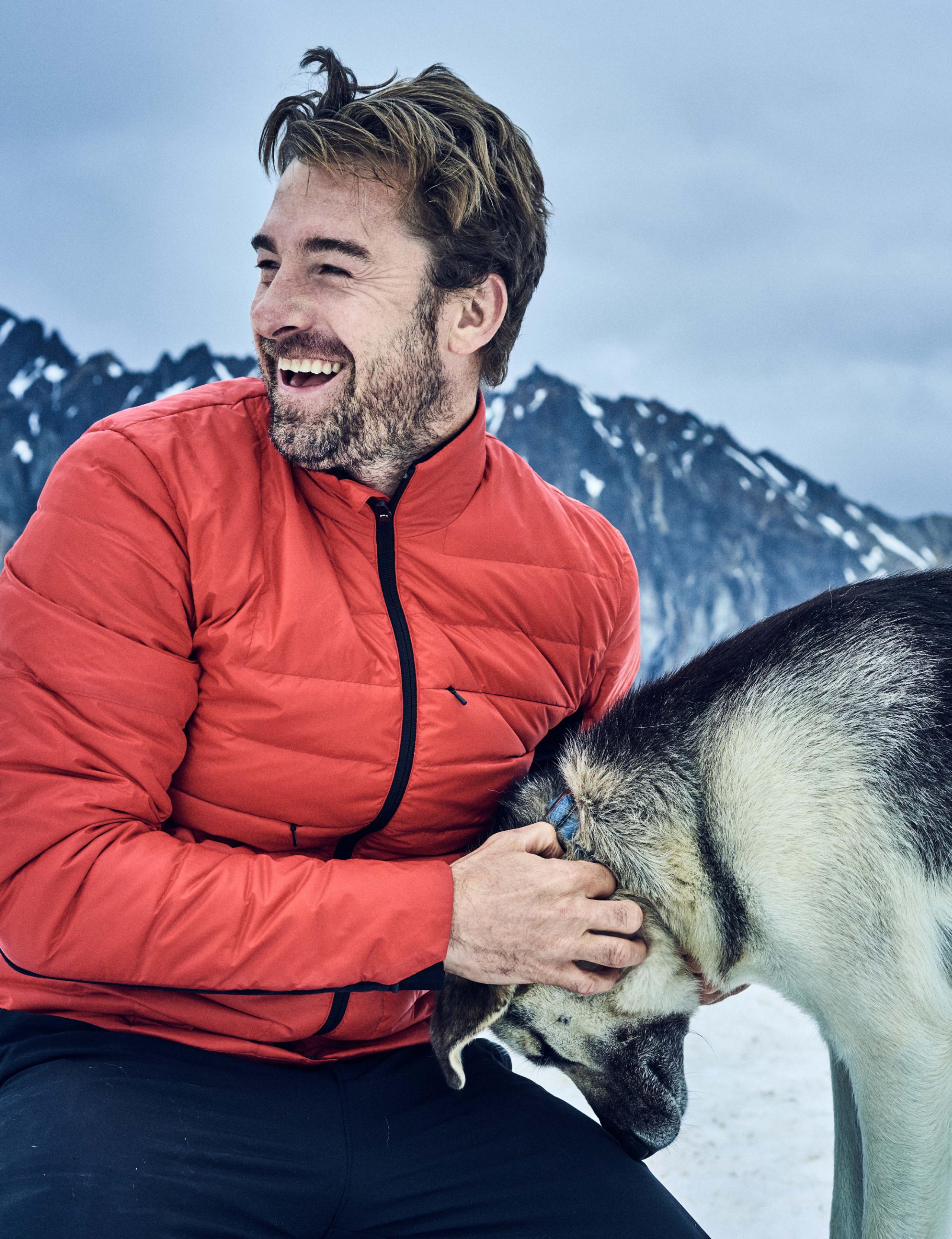 Man wearing red jacket smiling while scratching a dog on a glacier in Alaska