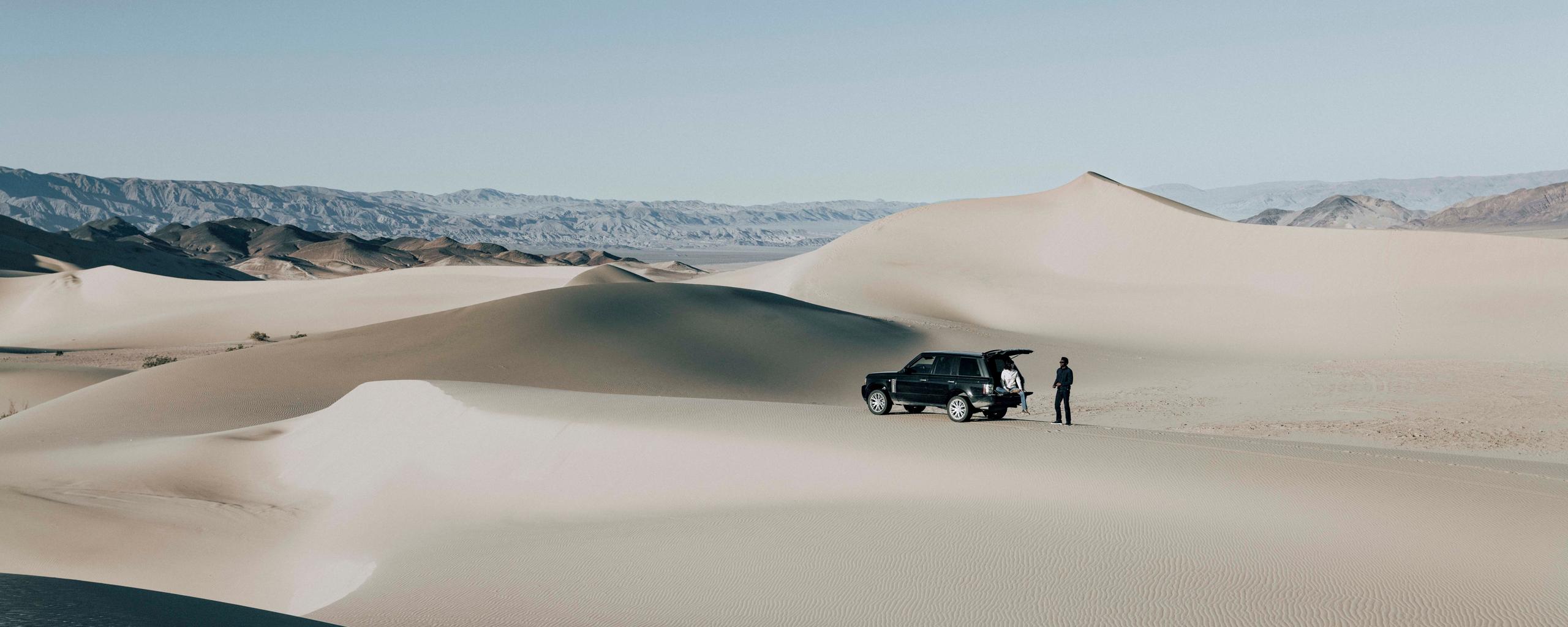 Landscape of desert sand dunes with man and woman outside Land Rover in the distance