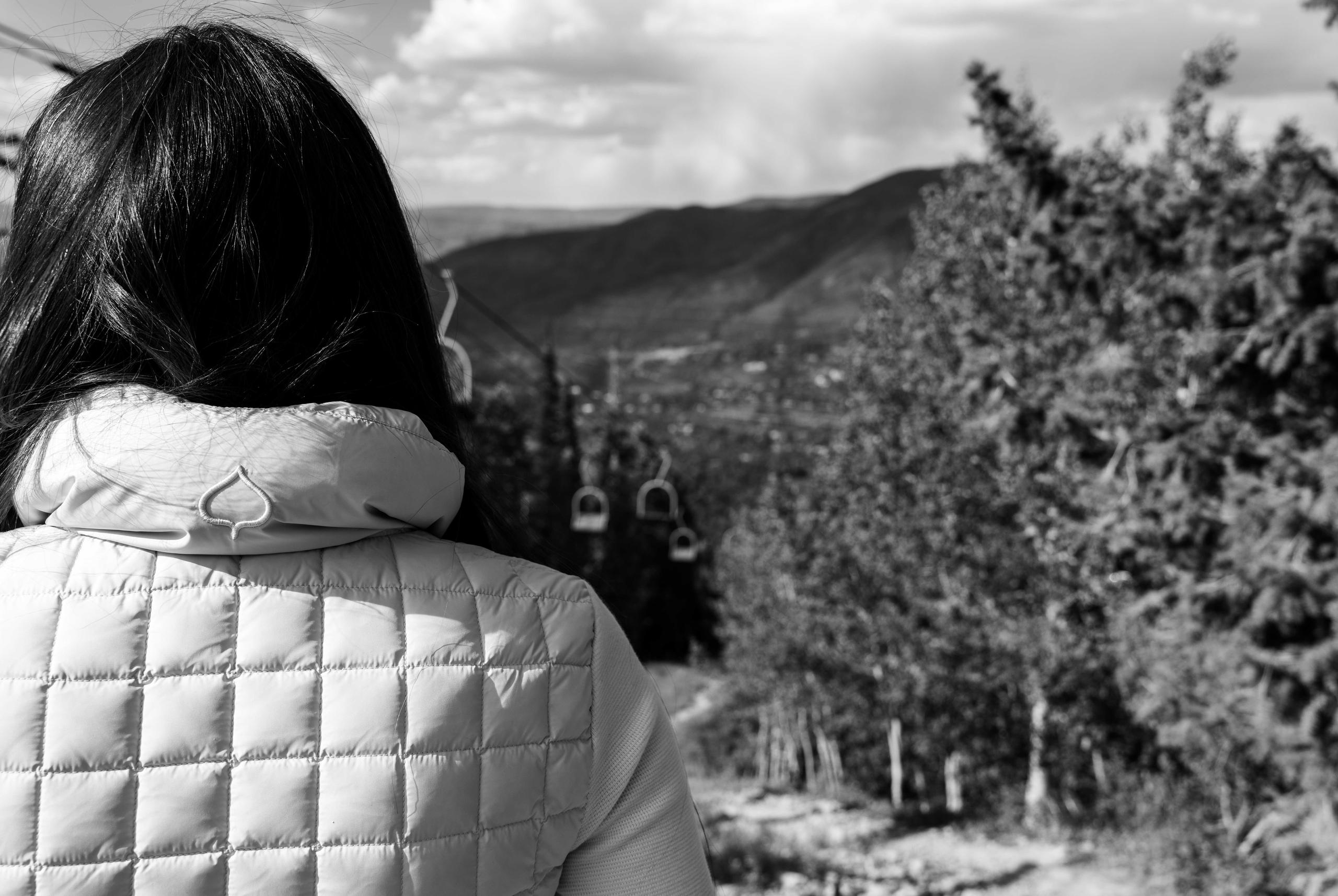 Black and white photo of back of woman facing Aspen ski lift and mountains in background