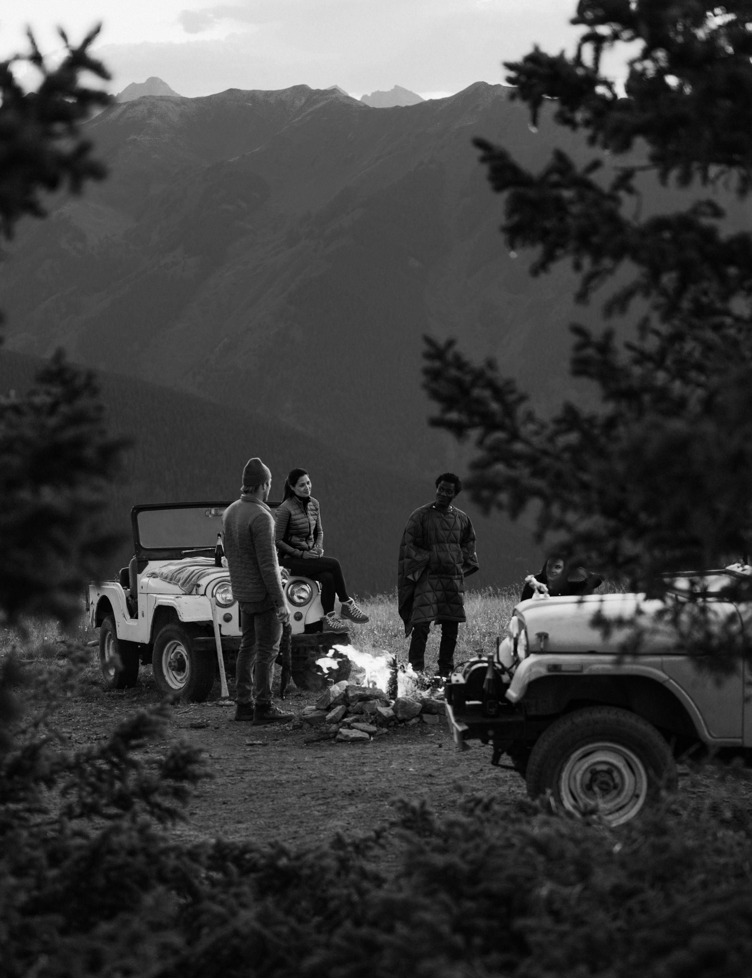 Pulled back black and white photo of two men and one woman sitting on vintage Jeep in front of campfire in Aspen mountains