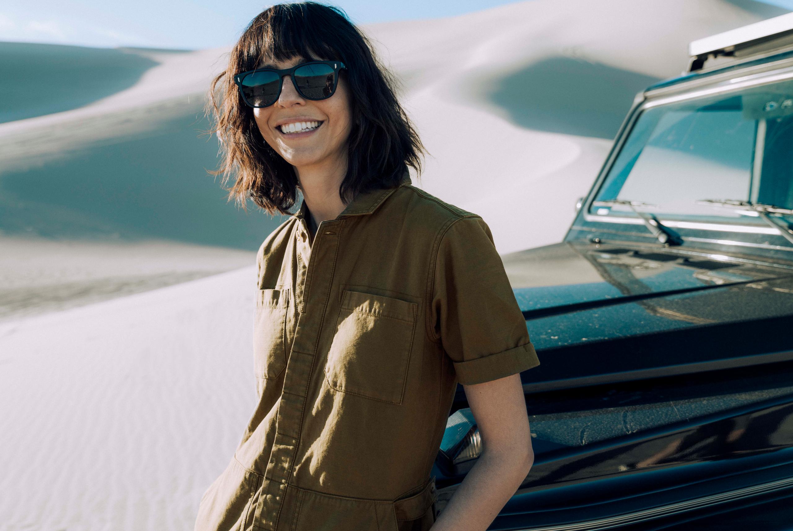 Woman wearing Yosemite sunglasses smiling in front of truck and sand dune background