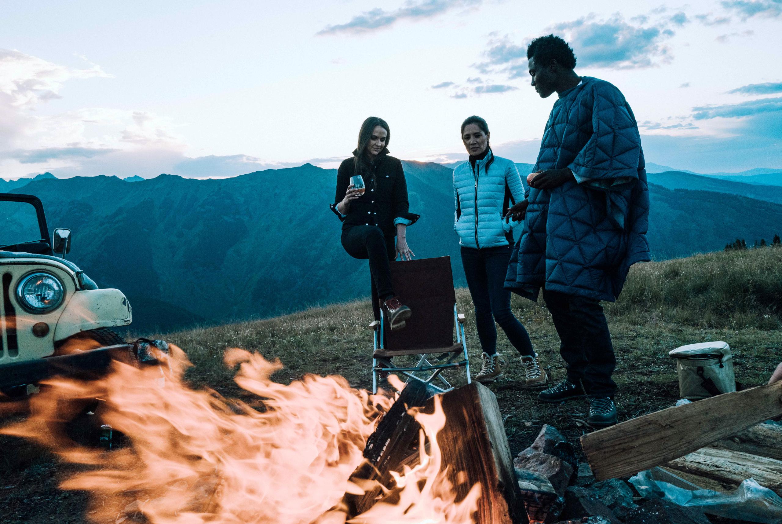 Two women and man standing around campfire alongside vintage Jeep in Aspen mountains