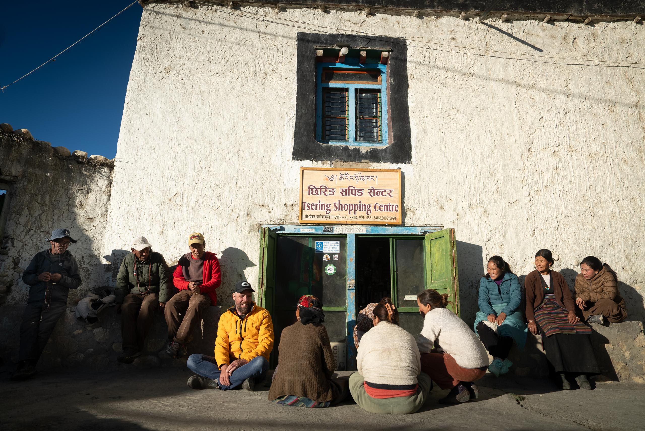 Traveler sitting on ground along with 9 locals in front of small shop in northern Nepal