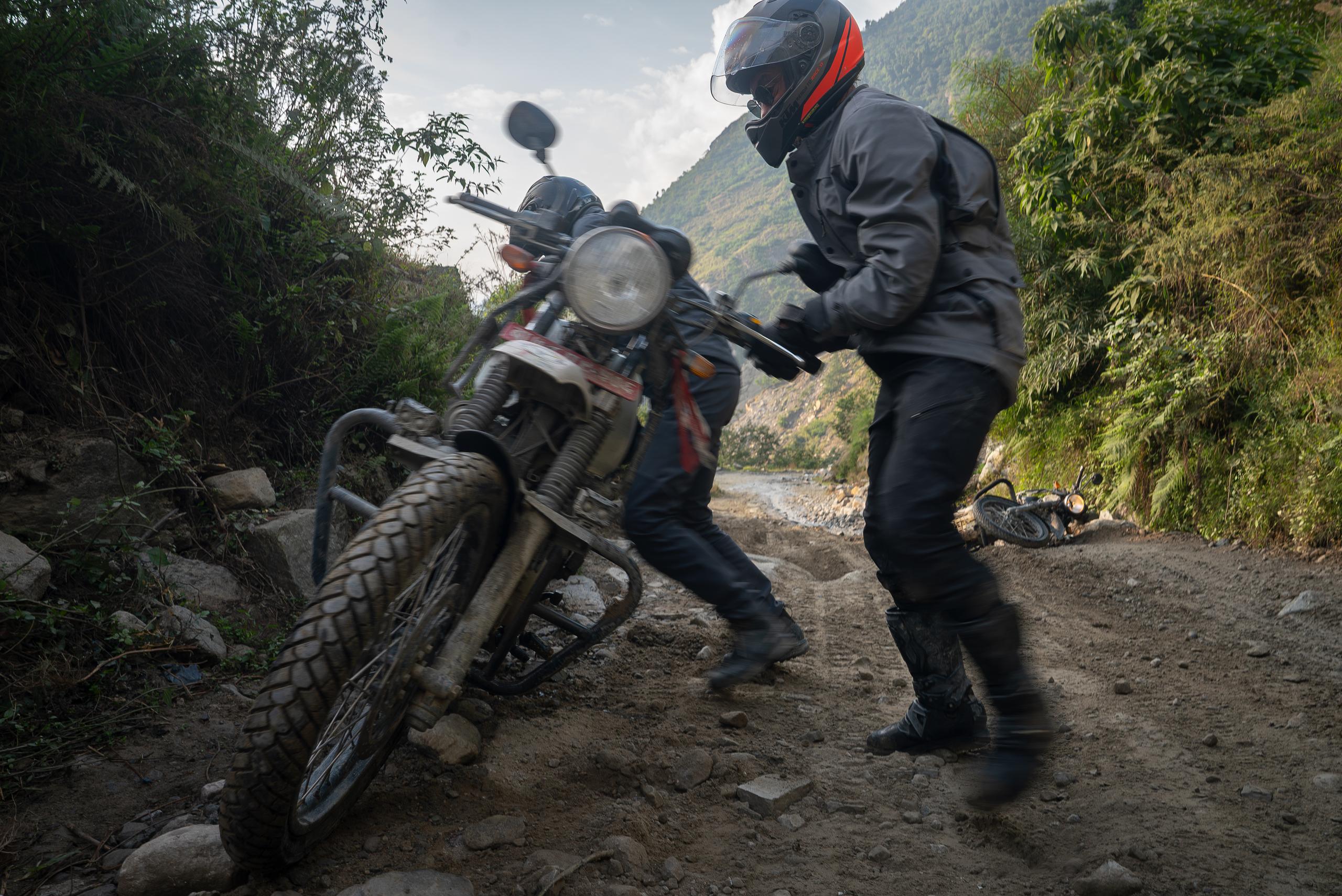 Two men lifting up fallen motorcycle on dirt road in mountains of Nepal