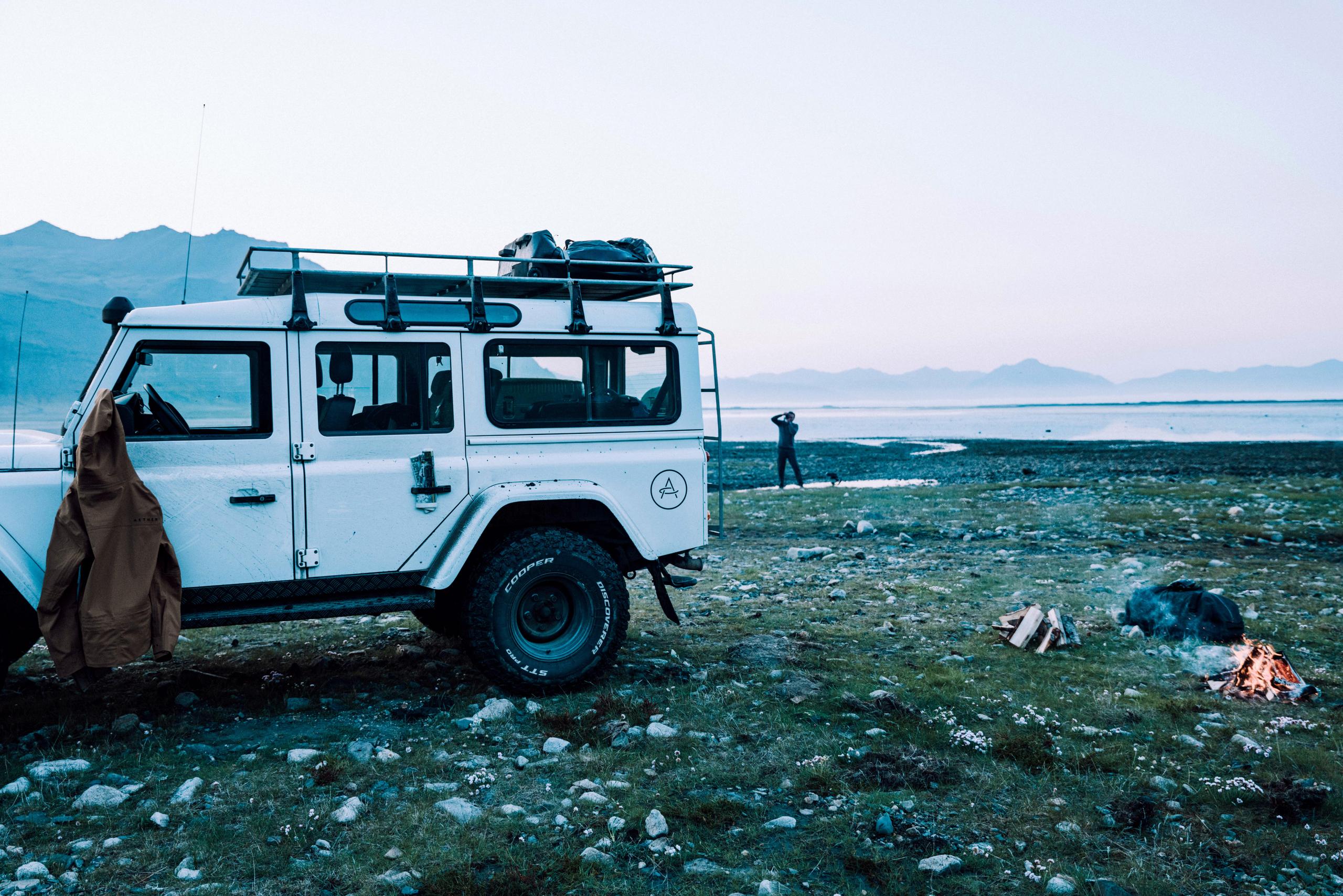 Camp site with white Defender, small campfire and man in the distance taking a photograph