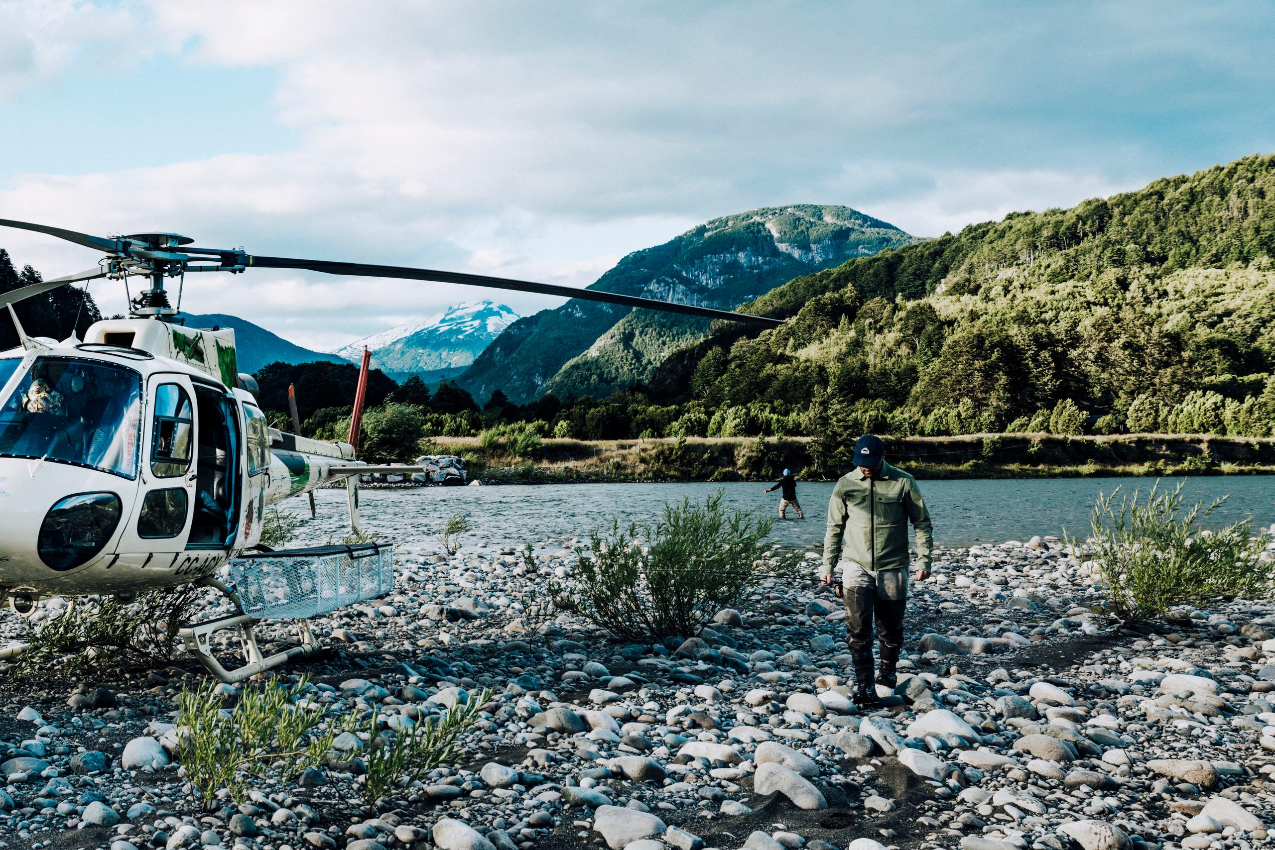 Man walking on rocky shore alongside Patagonia river with helicopter parked nearby