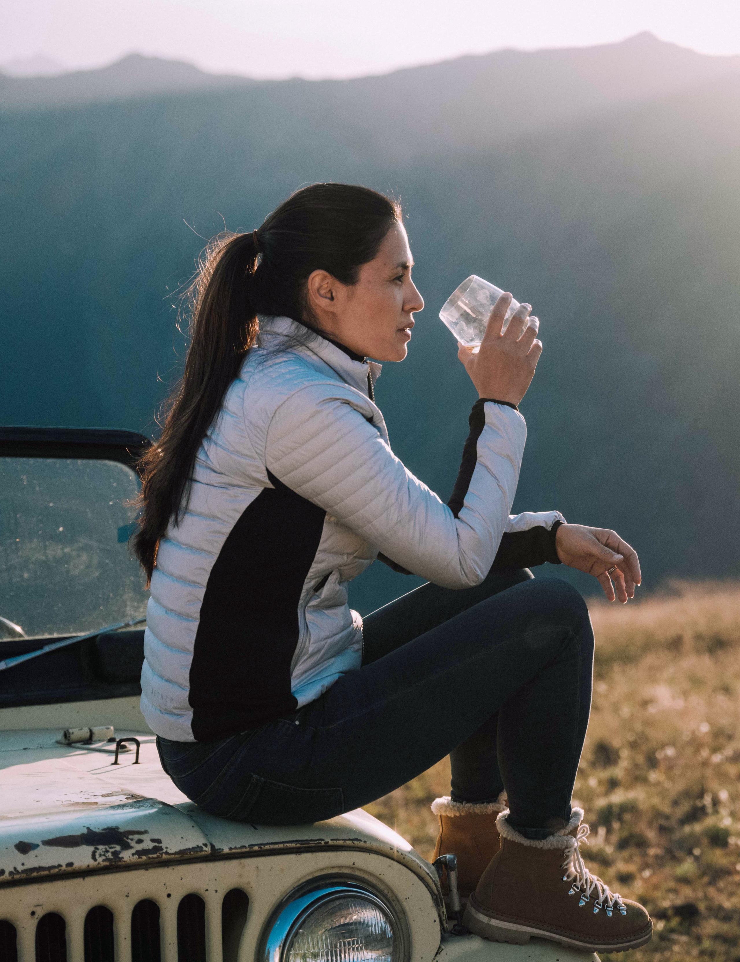 Woman drinking from cup sitting on top of vintage Jeep in front of Aspen mountains