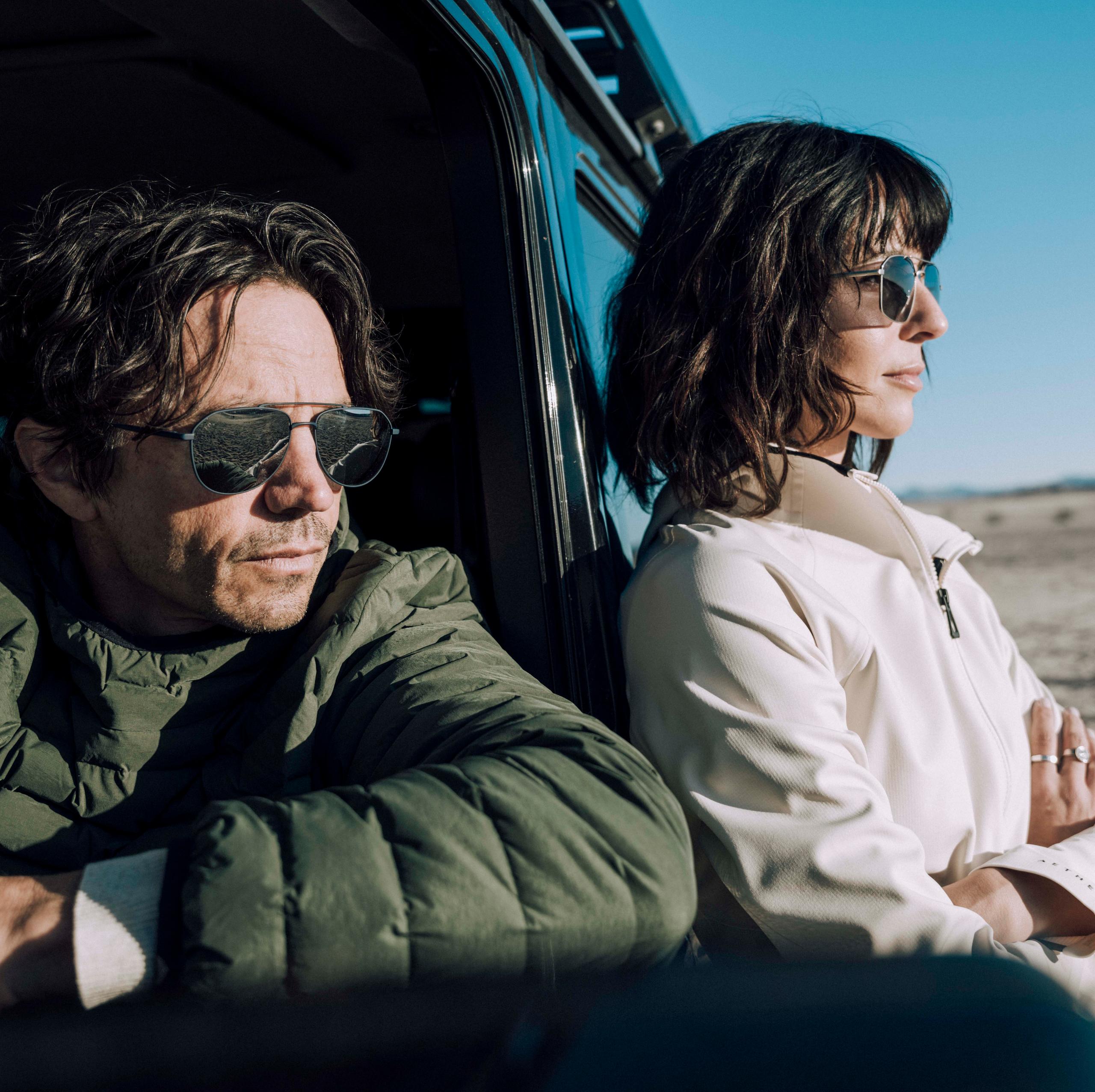Man in driver seat and woman leaning against car looking out to vast sand dunes