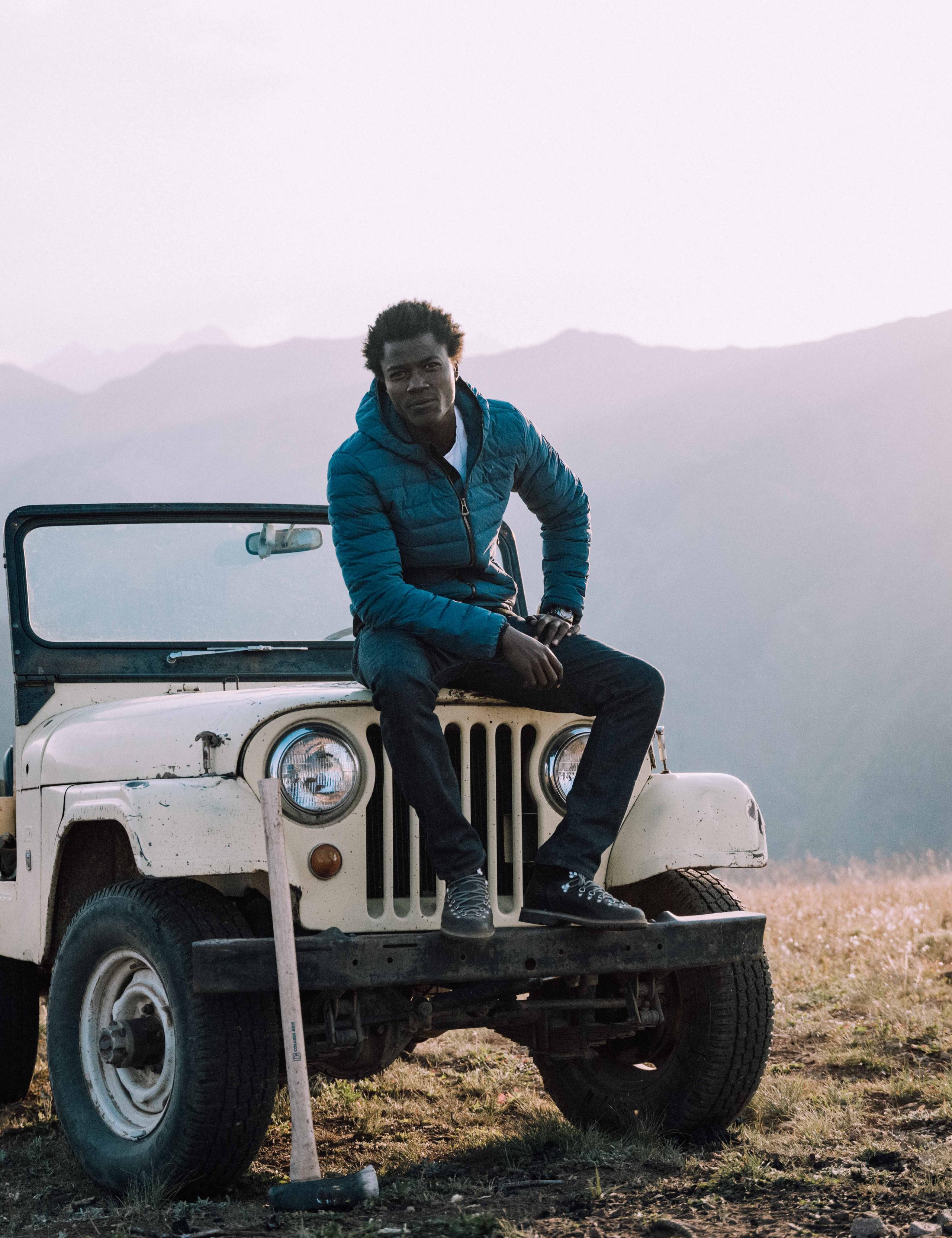 Man standing on front bumper of vintage Jeep in field overlooking Aspen mountains and sunset  