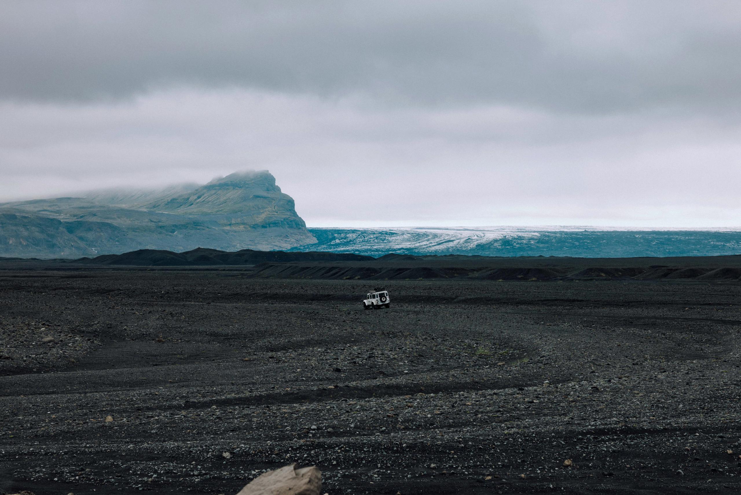 Small white Defender in the distance of black rocky terrain in front of glacier in Iceland