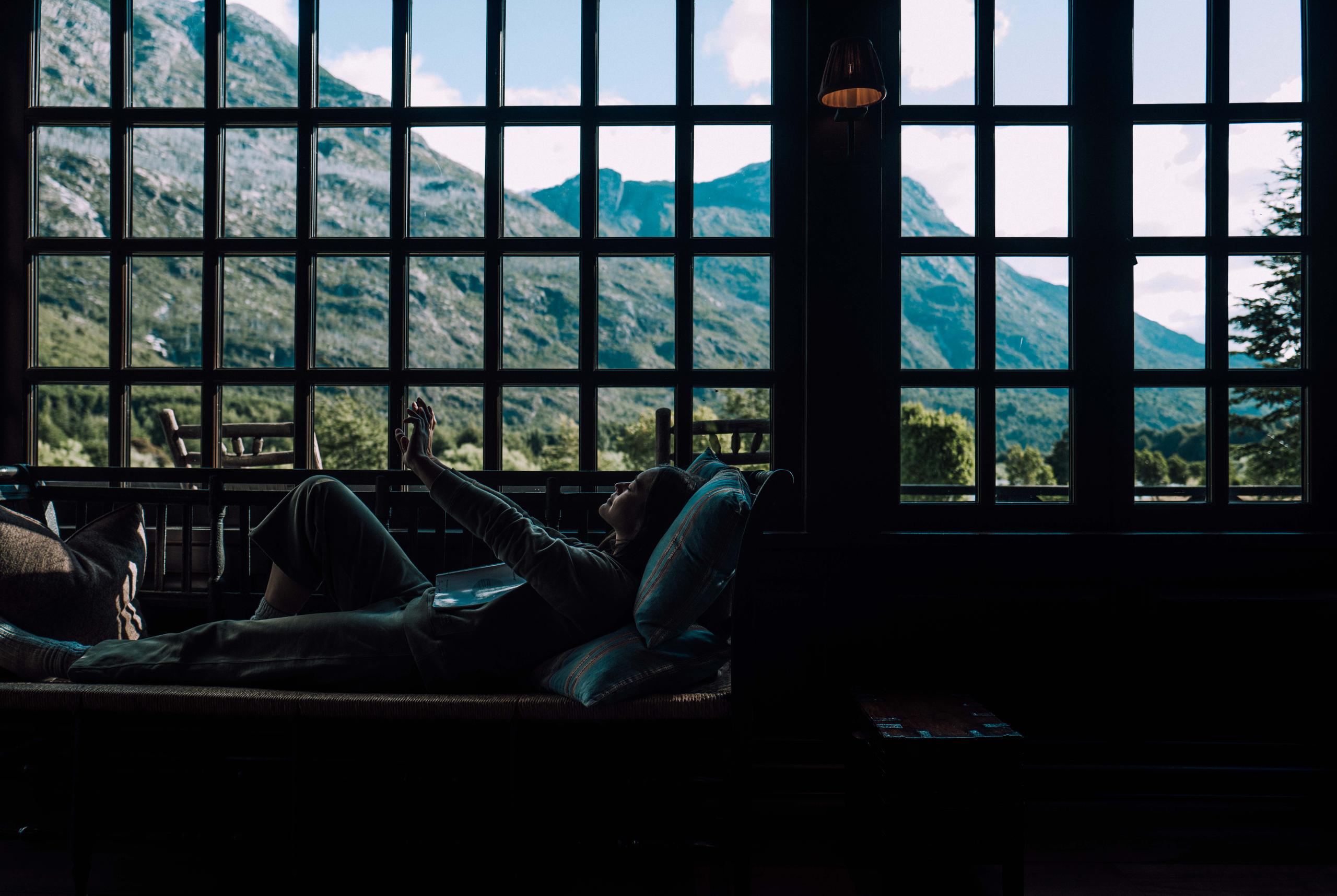 Woman lounging inside Eleven Lodge next to large window overlooking mountains of Patagonia