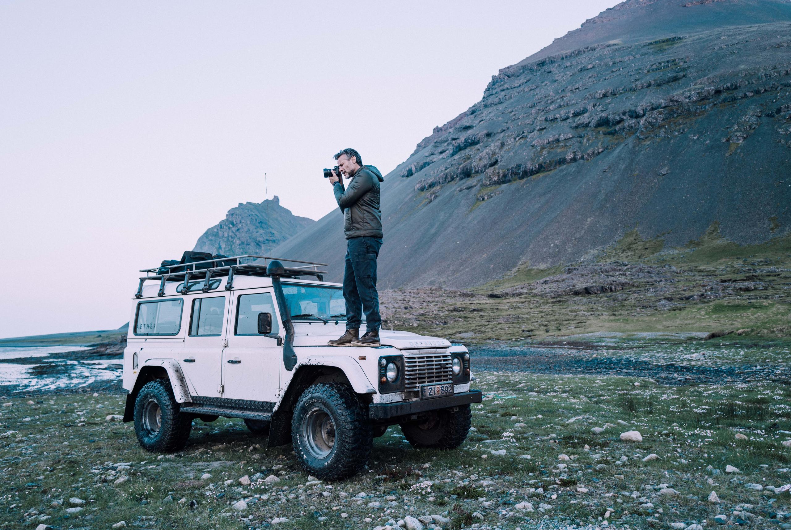 Man with camera standing on top of white Defender truck in Iceland landscape