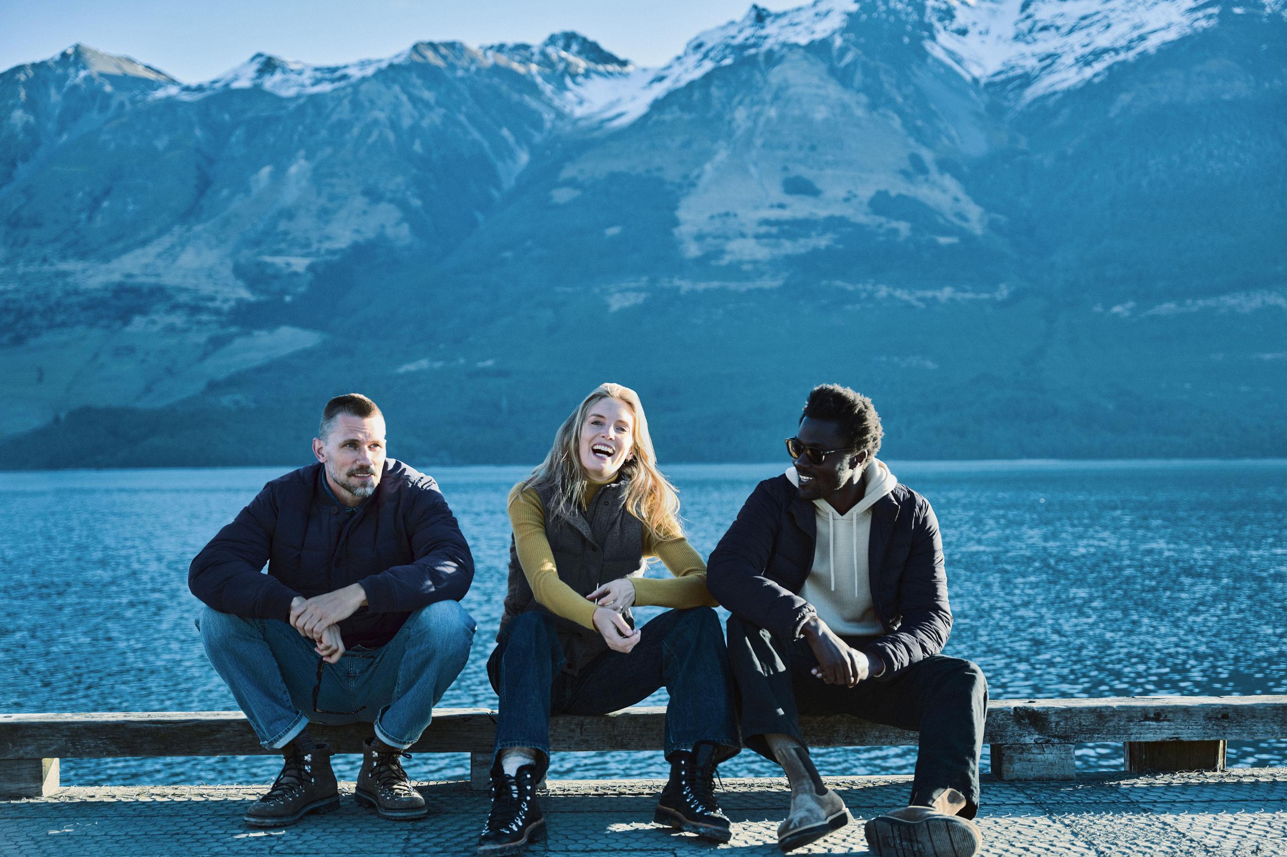 Three people sitting on an iron bridge over a lake in New Zealand laughing pleasurably.