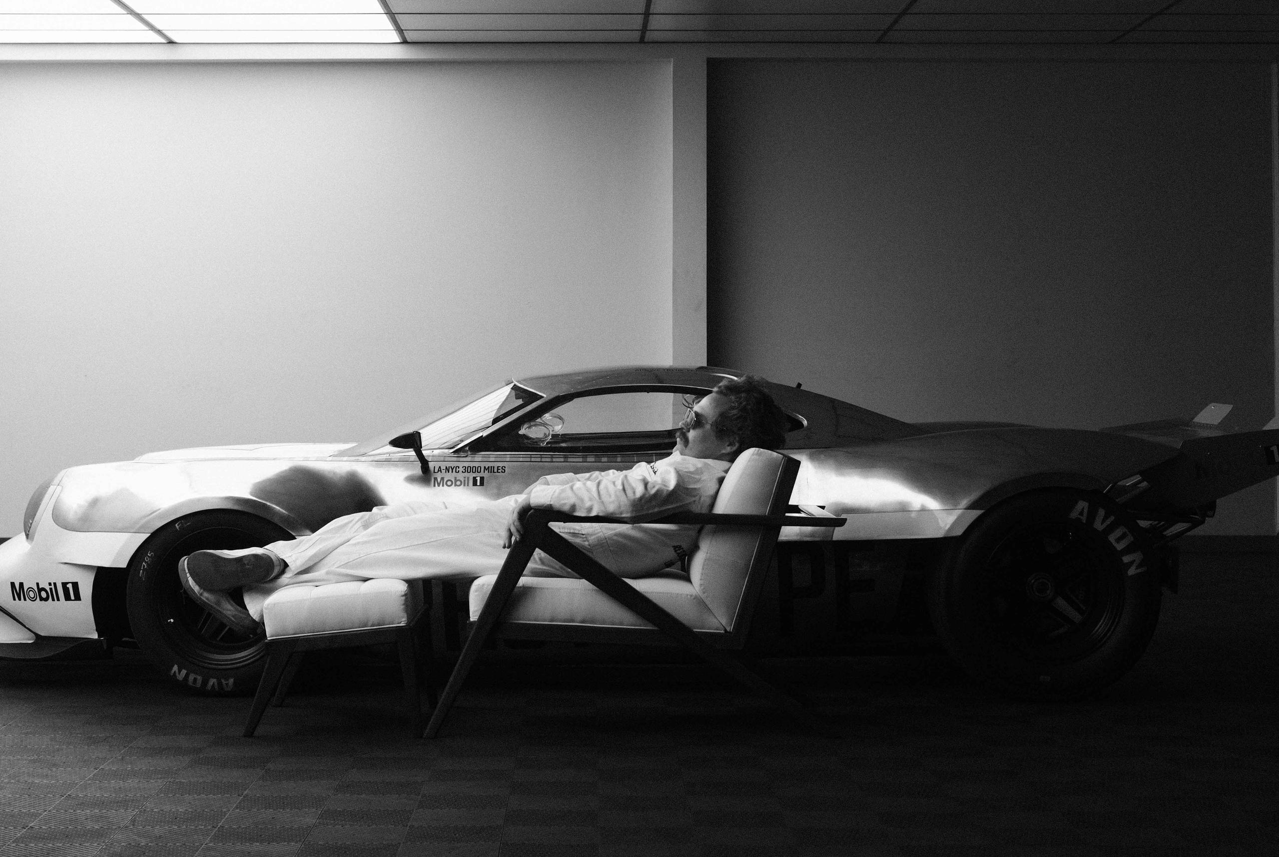 Black and white photo of Nikita Bridan lounging on chair next to the Half11 race car prototype