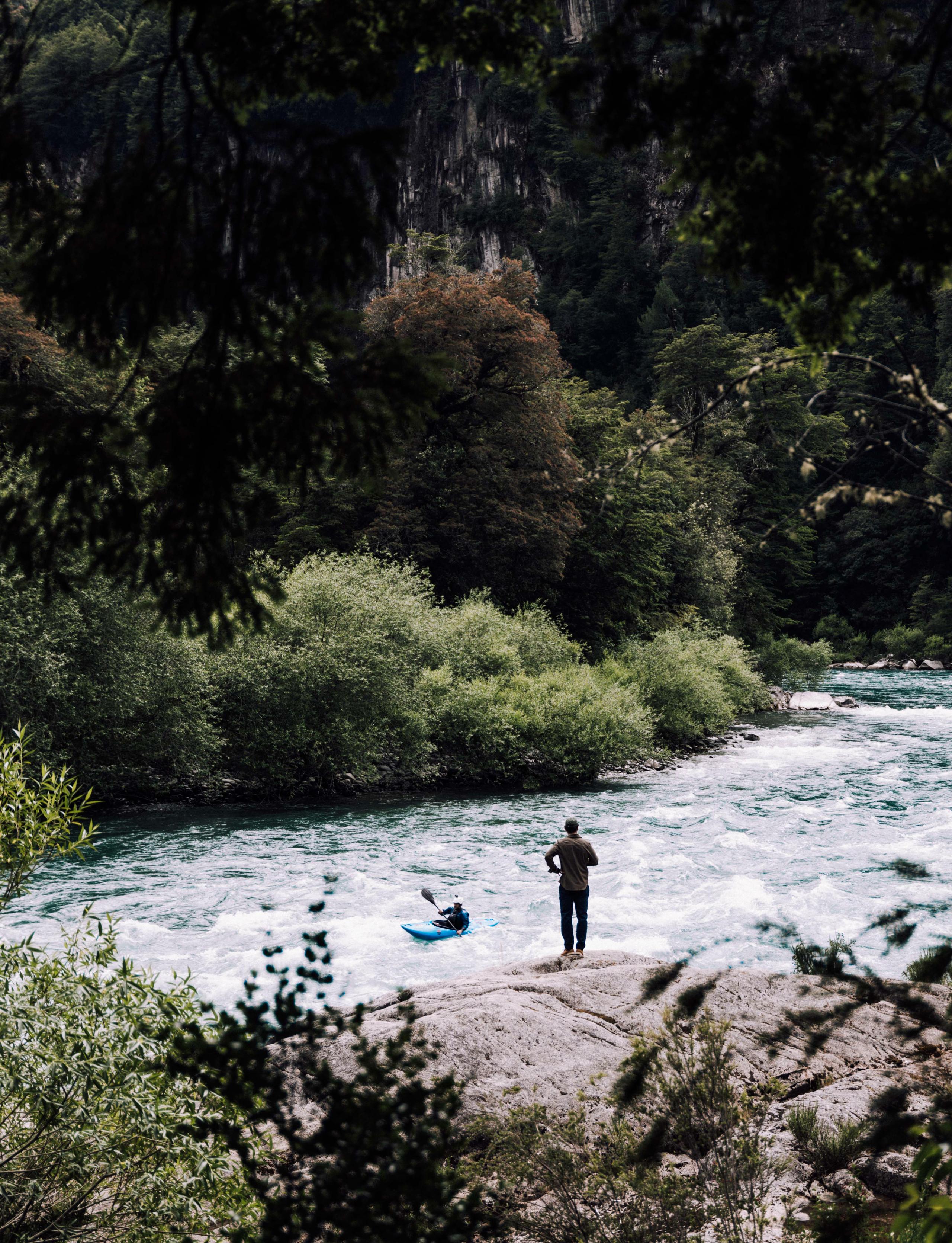 Man on edge of river watching person kayaking down river in Patagonia