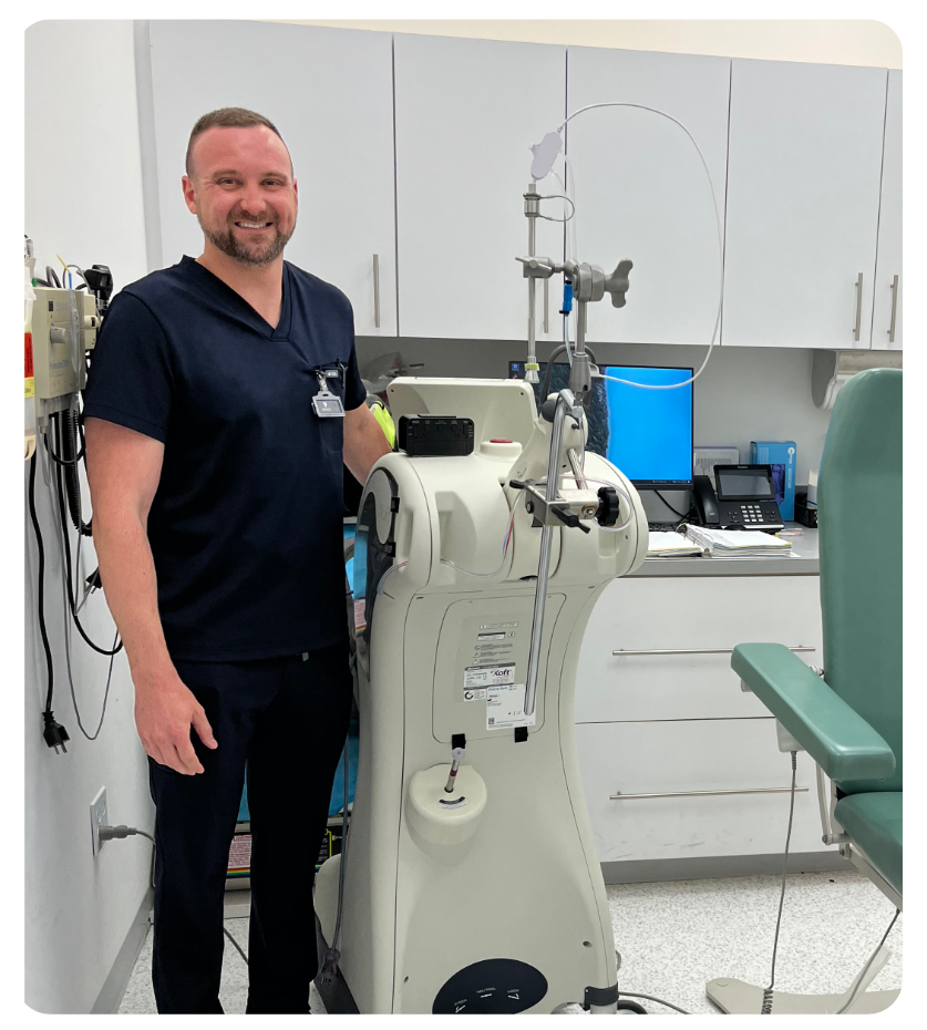 Joe Strahl in navy scrubs, standing beside the brachytherapy unit in a Sunnycoast Dermatology treatment room.