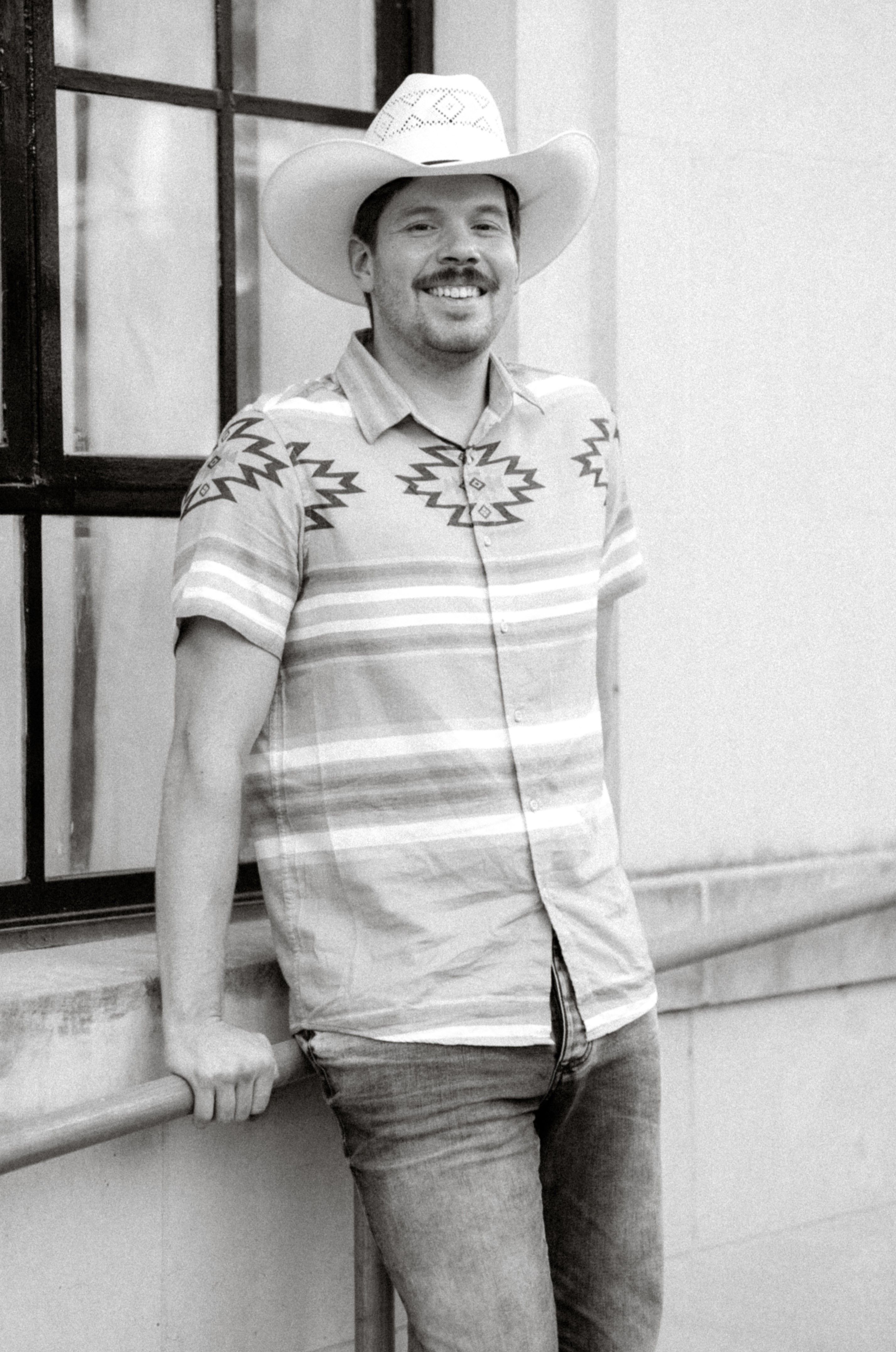 A black and white photo of a man with a cowboy hat leaning on a railing