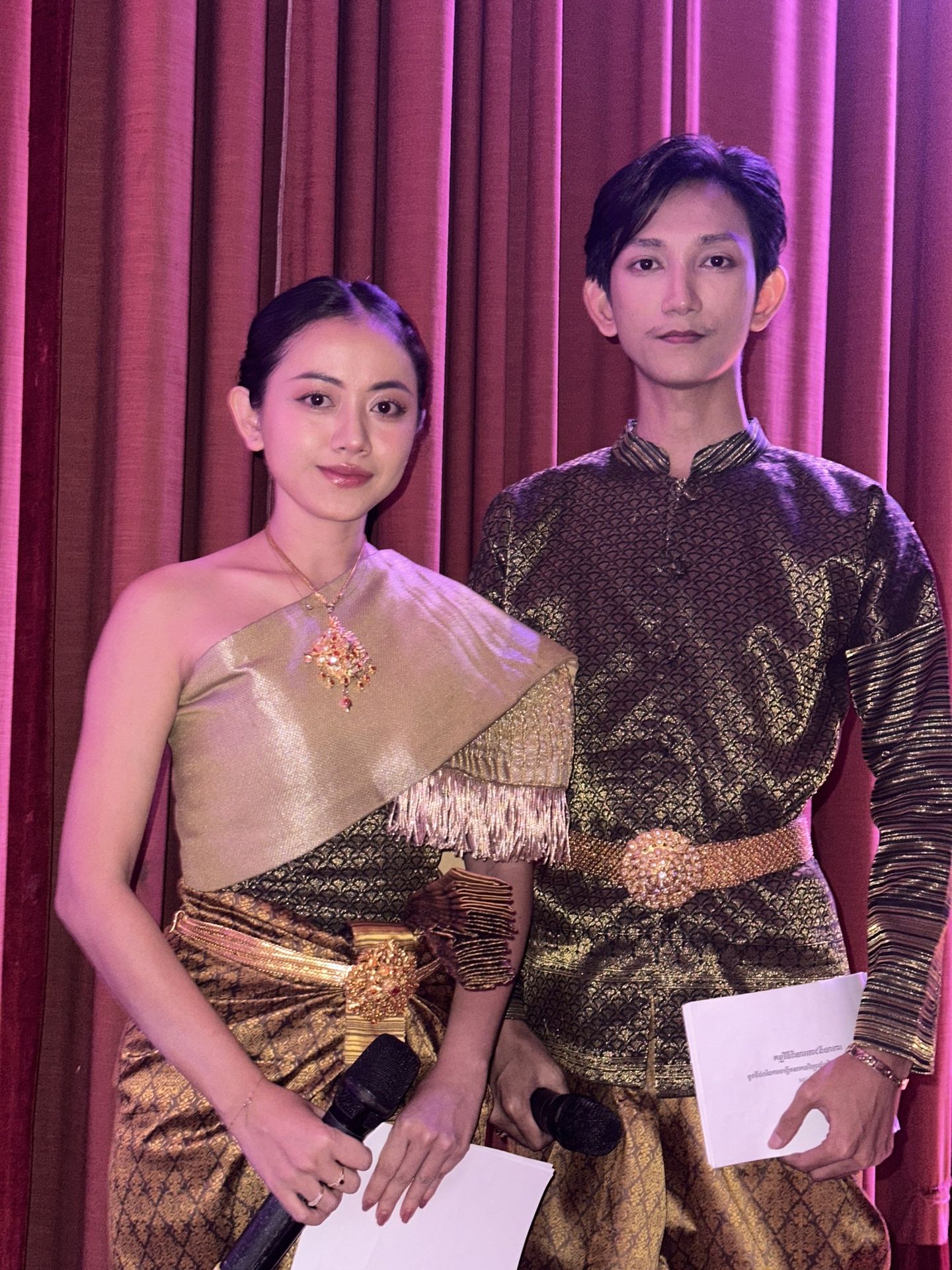 Serei Van Kosaun and her fellow MC in traditional Khmer dress before a crimson curtain: she in gold with a draped shoulder cloth, he in a patterned jacket and gold belt
