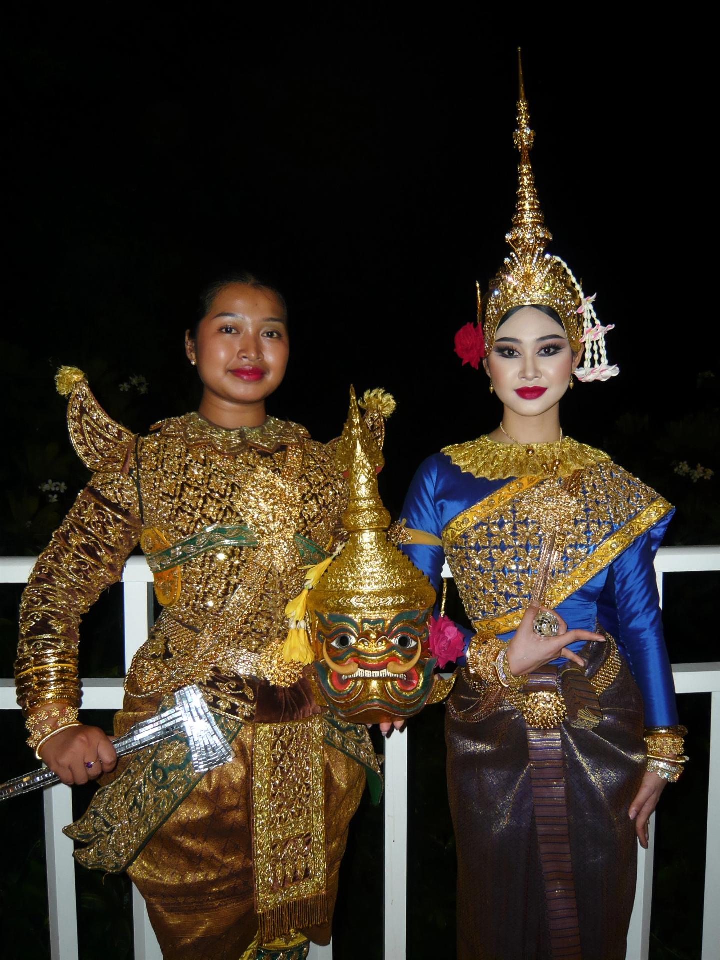 Two performers in ornate Khmer costumes pose together; the performer on the left holds a giant mask and axe, while the performer on the right wears blue and gold with a tall crown.
